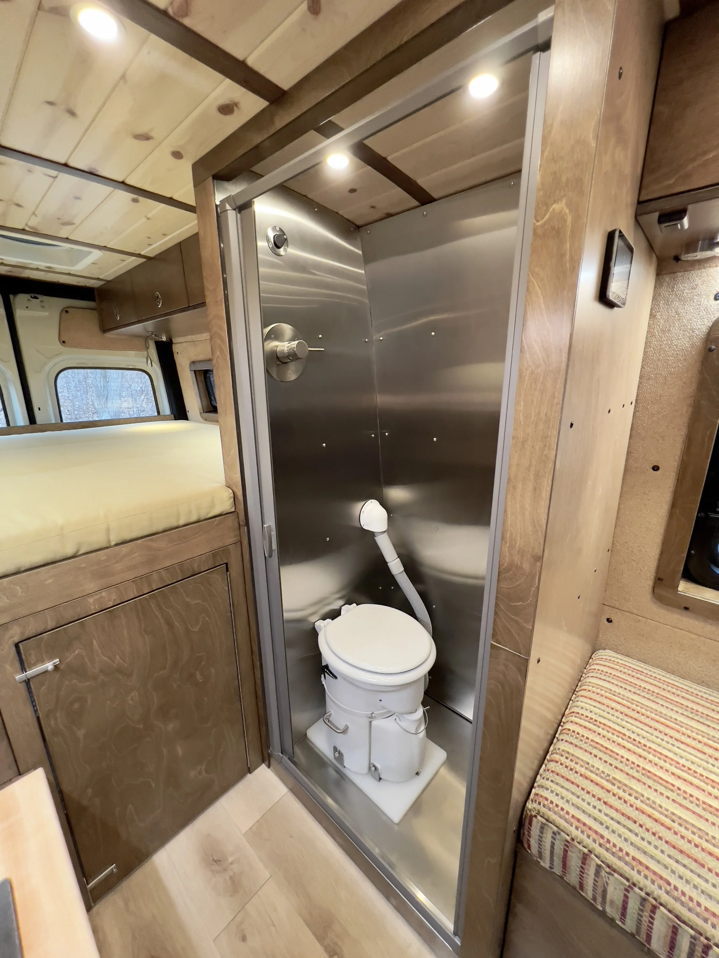 Small bathroom with a portable toilet inside a shower enclosure, wooden walls, and a bed with a yellow mattress nearby.