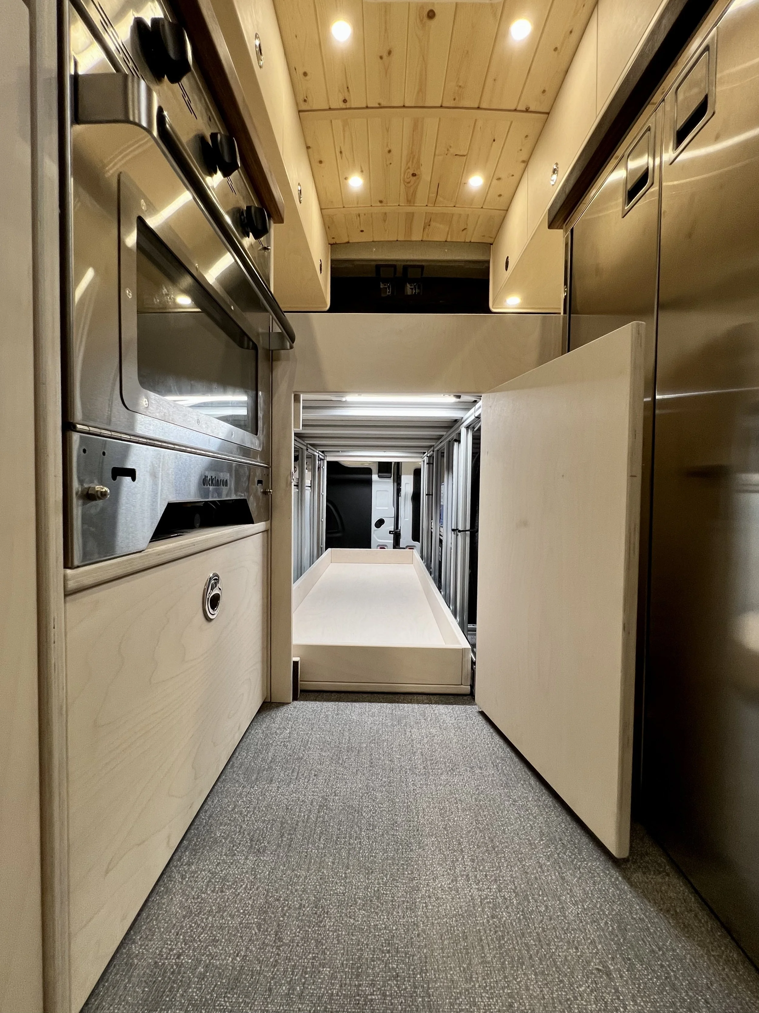 Interior view of a compact kitchen and living area in a converted camper van, featuring a wood-paneled ceiling, stainless steel appliances, and a low bed platform at the back.