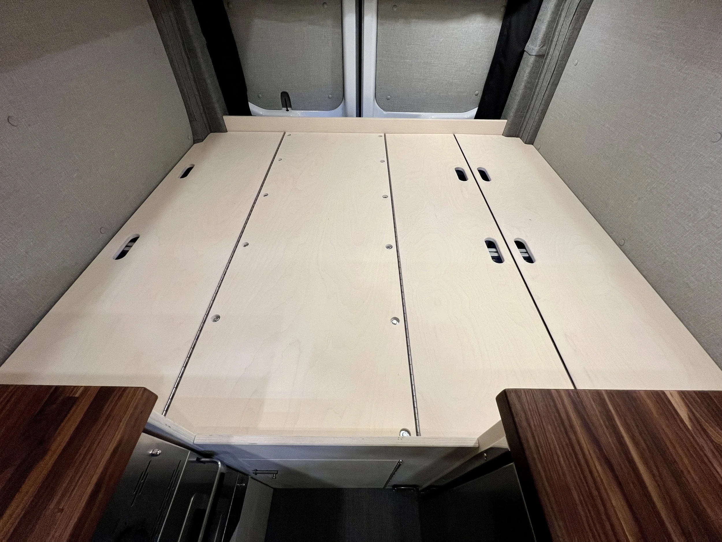 Interior of a vehicle with the back seats folded down to create a flat surface, possibly for a bed or storage, with tan walls and wooden tables on both sides.