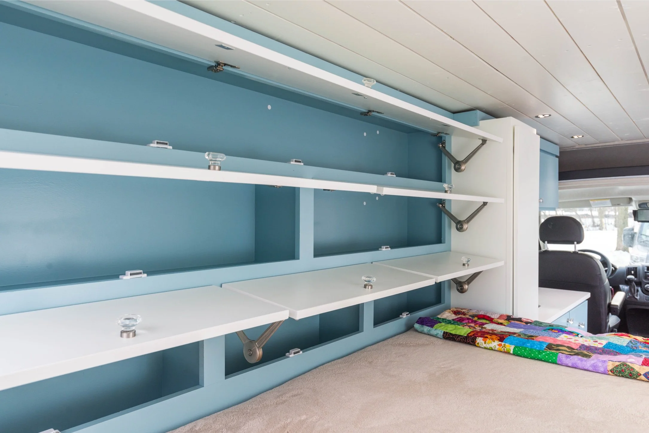 Interior of a van with a custom blue shelving unit, white fold-out tables, and a beige bed with colorful quilt, driver and passenger seats visible in the front.