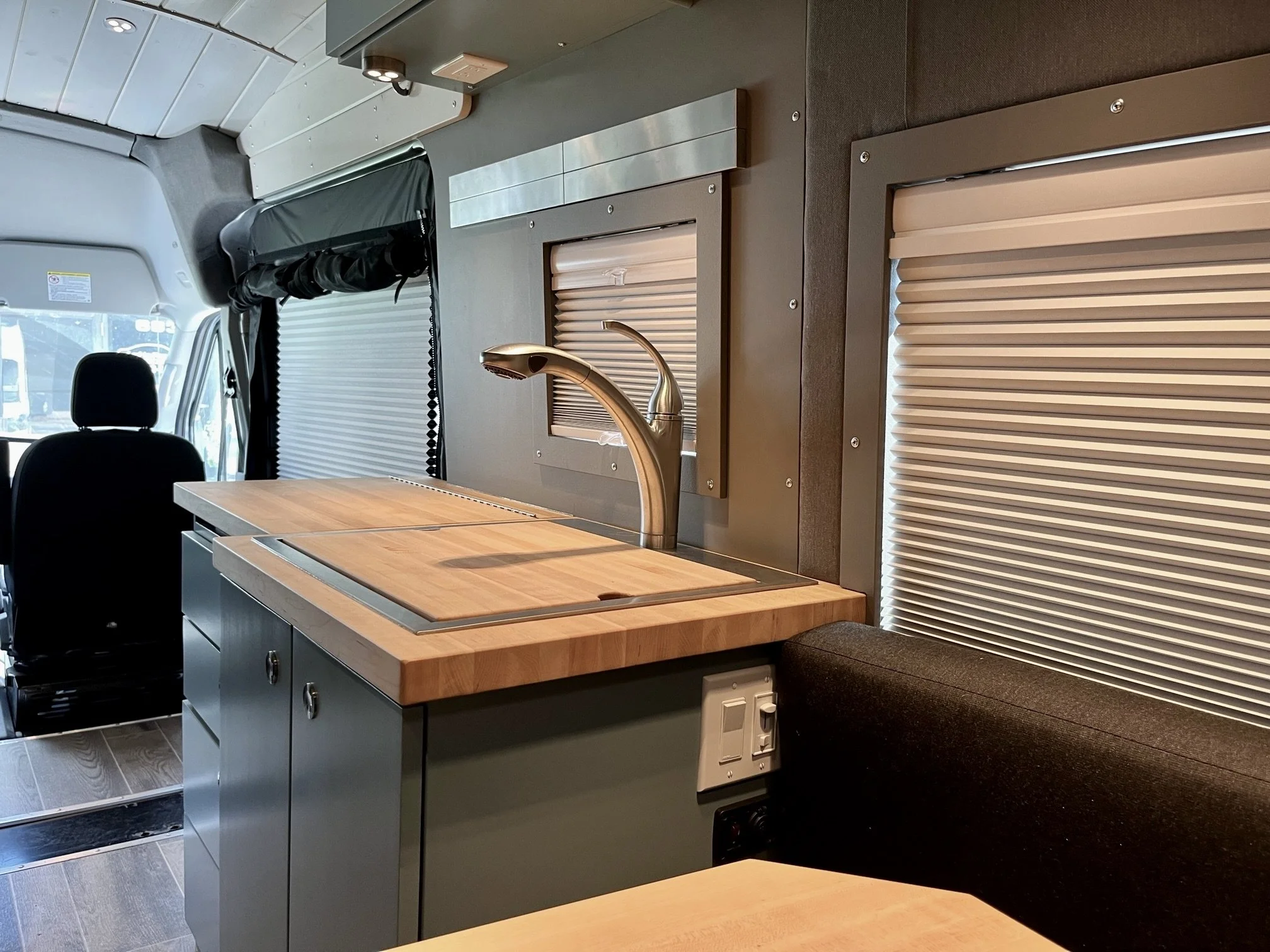 Interior of a camper van featuring a wooden countertop with a built-in sink, a sleek brass faucet, and closed wooden cabinet doors underneath. There are two windows with beige blinds, and the front cabin with a driver's seat is visible in the background.