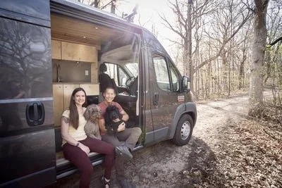 Two women and a man sitting outside a gray van, smiling, with one holding a small black dog, in a wooded area.