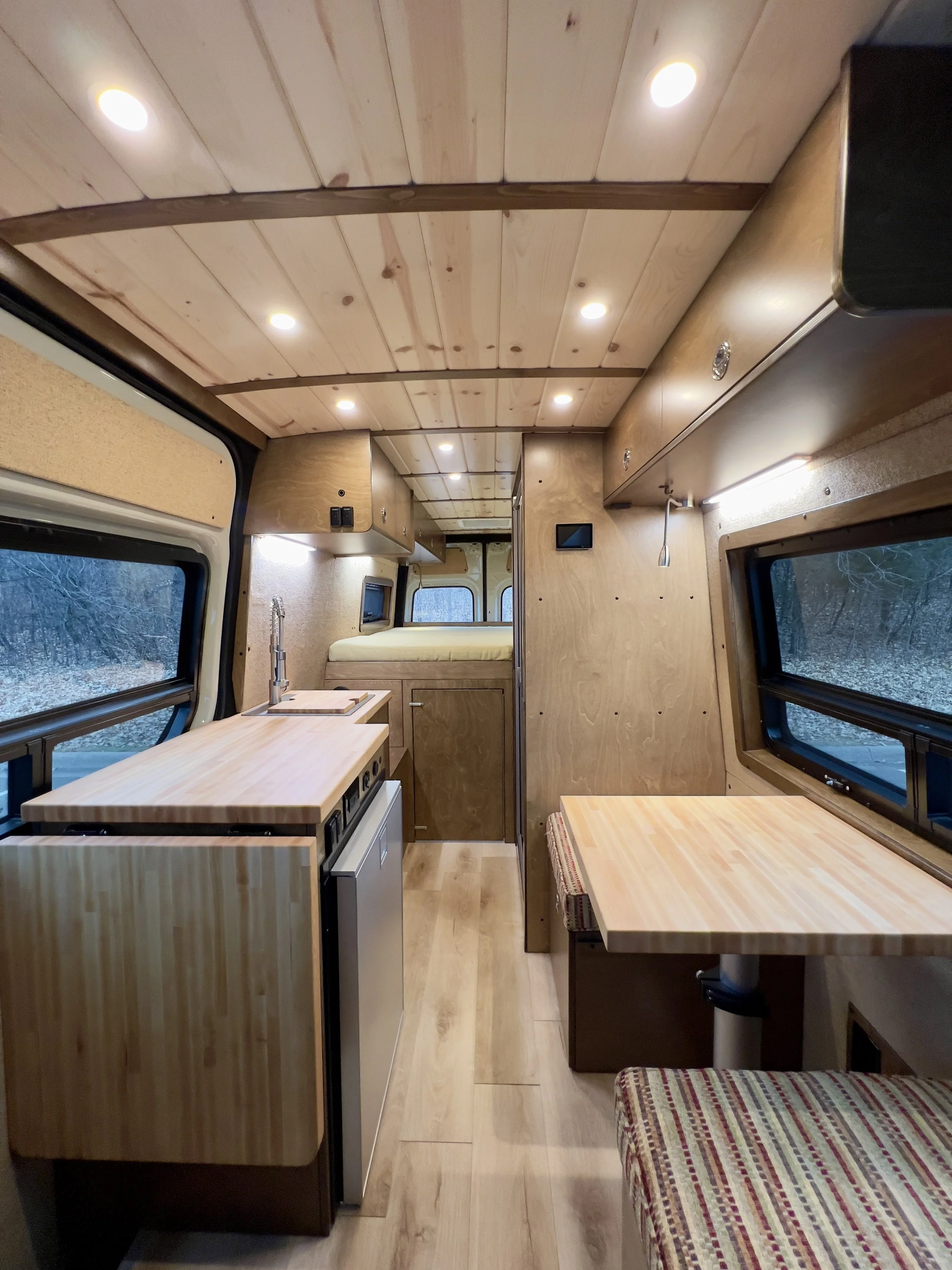 Interior of a tiny camper or van converted into a living space with wooden cabinetry, a small kitchenette with a sink, a seating area with a table, large windows, and a sleeping area in the back.