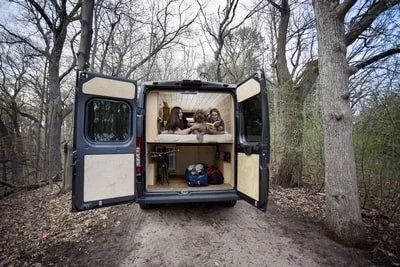 Three people sitting at a table inside the back of an open van on a woodland dirt trail, with trees surrounding them.