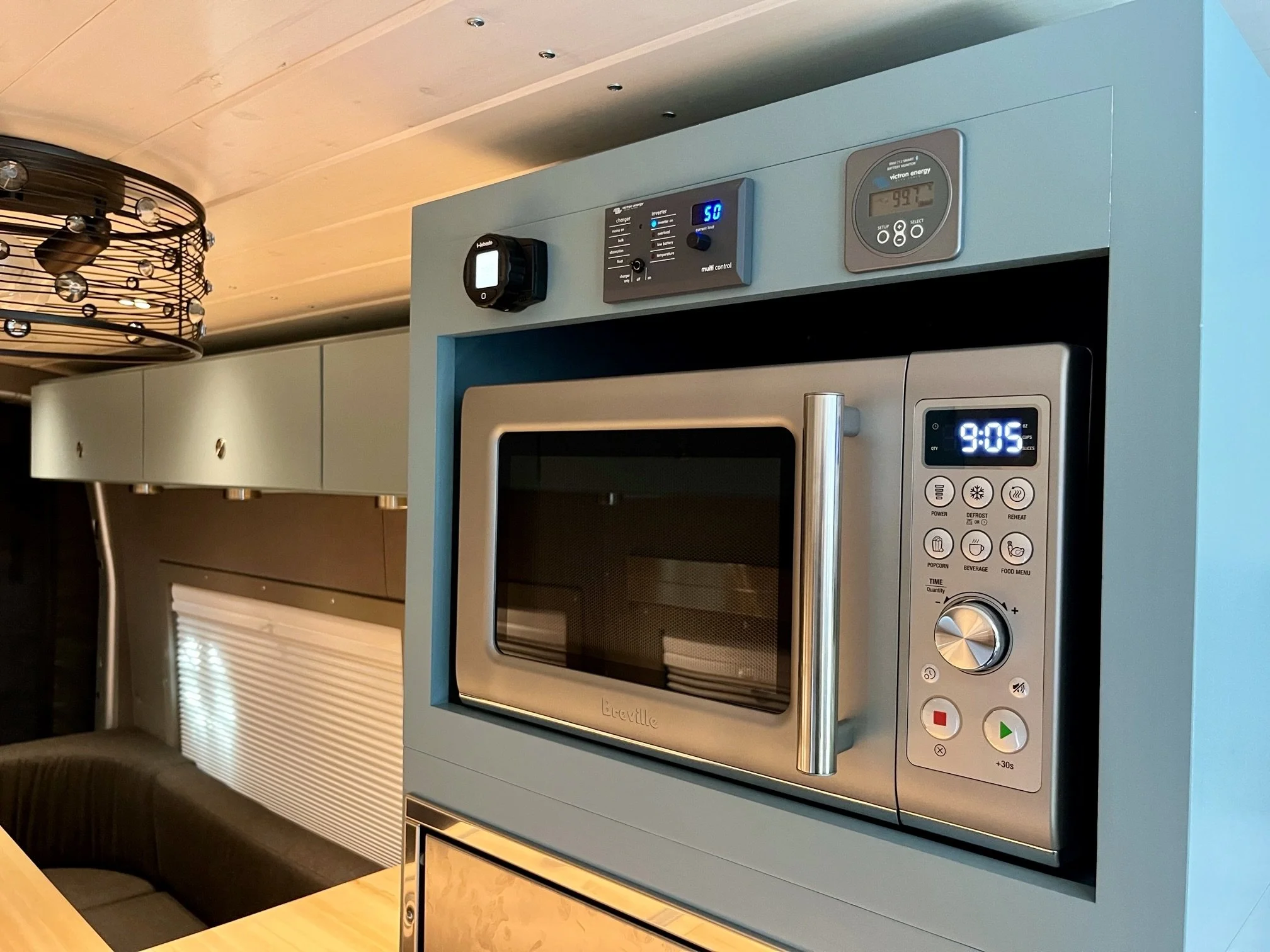 A microwave oven built into a kitchen wall with digital display showing 9:05, various control buttons, and a metallic handle. Above it are additional electronic controls and displays.