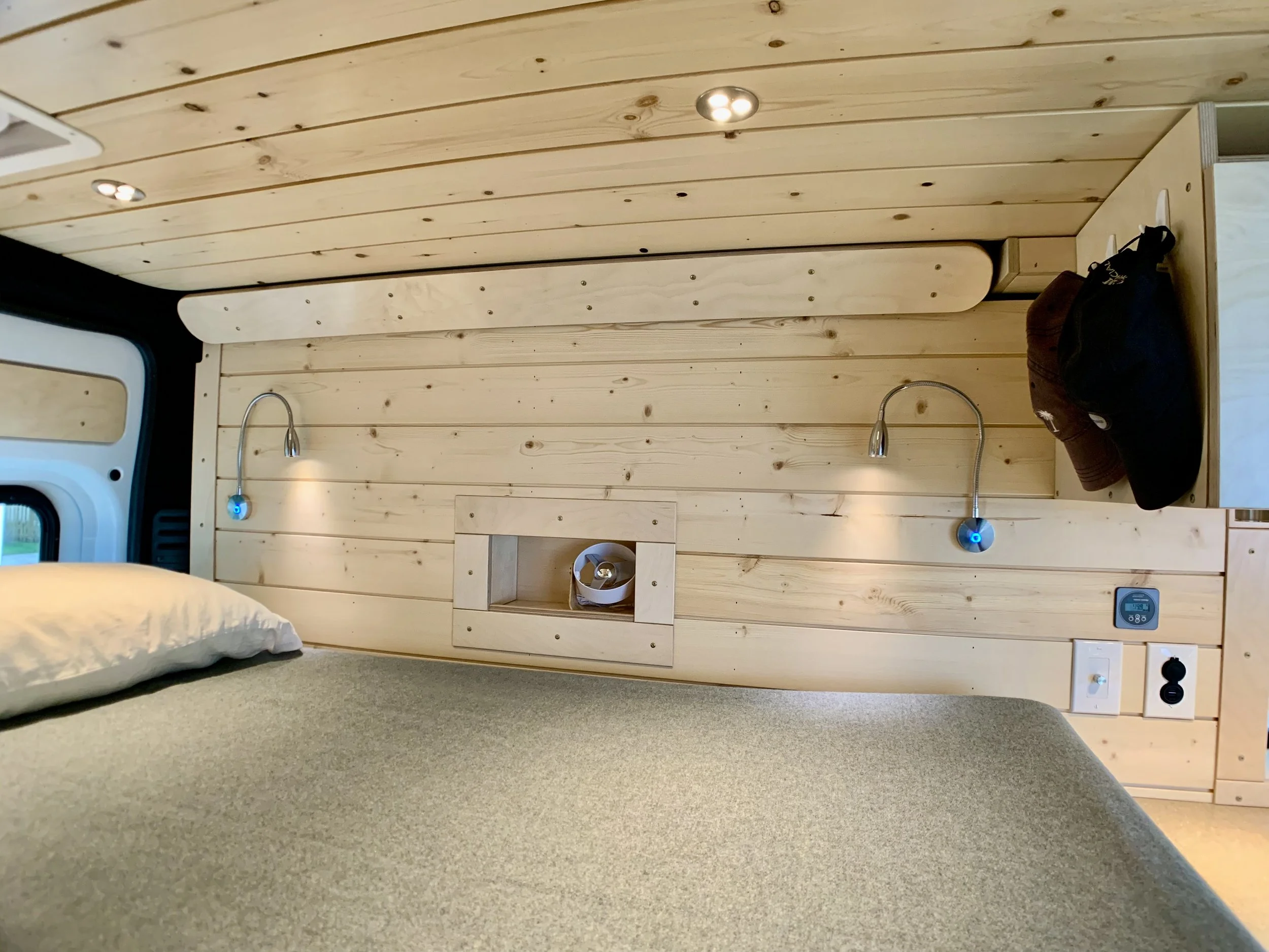 Interior view of a small wooden bedroom with a bed in the foreground, two adjustable reading lights, a small built-in shelf, and hooks with hats and clothing on the wall.