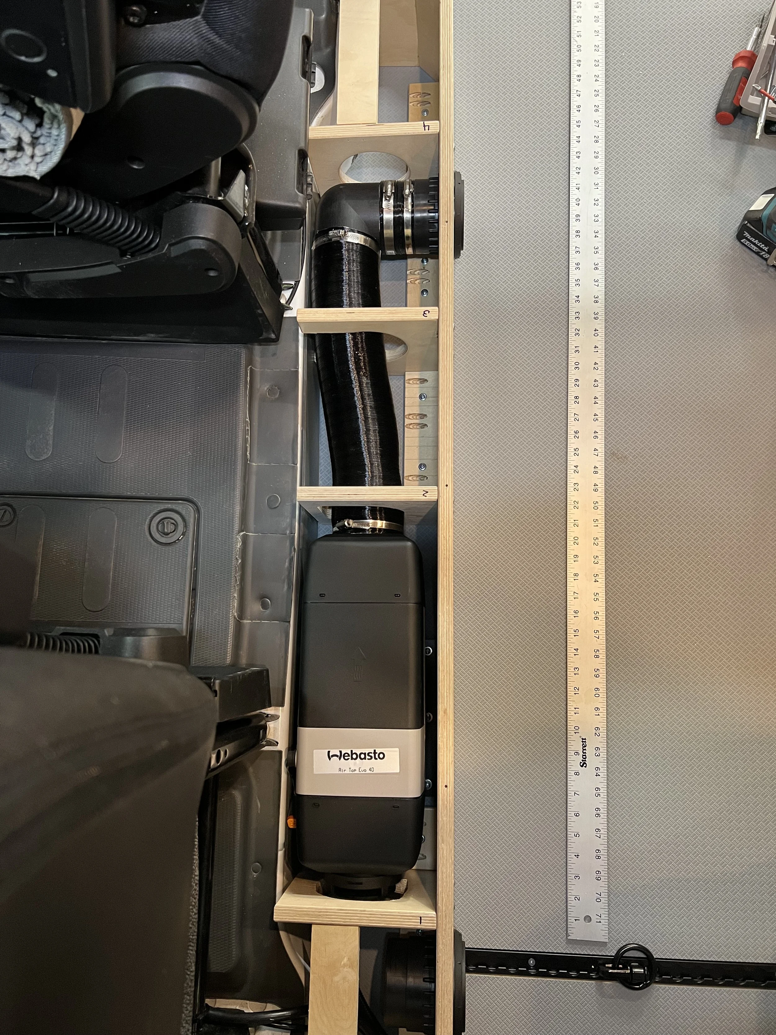 A measuring system installed in a workshop setup, featuring a wooden frame, a black air compressor labeled 'Webasto,' and a large ruler for measurement.