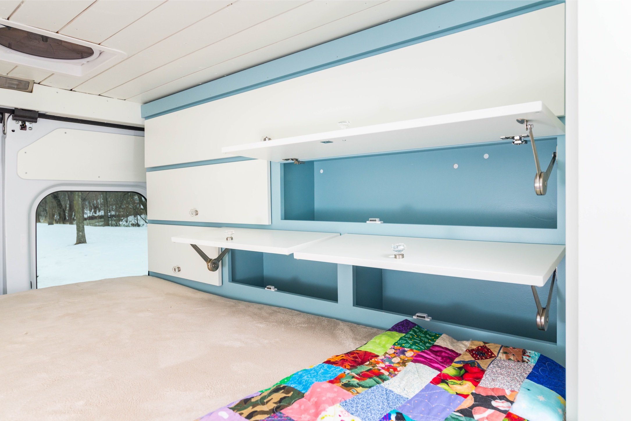 Inside a camper van with a bed, multi-colored quilt, and built-in storage compartments with white and blue surfaces