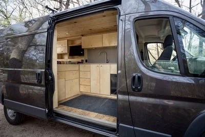 Interior of a converted camper van with wooden cabinets and shelves, gray flooring, and a small flat-screen TV.