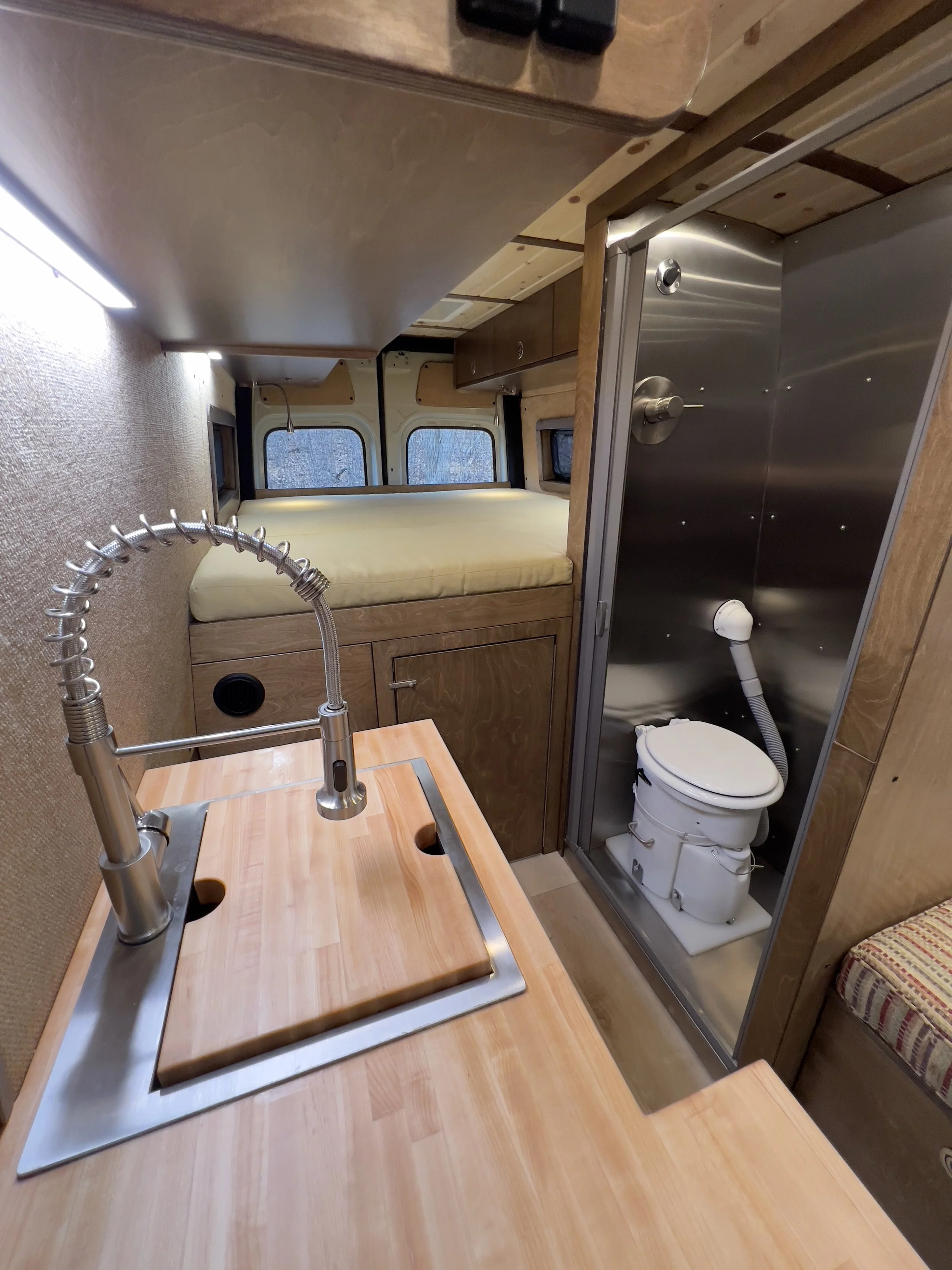 Interior of a compact camper van, showing a small kitchen sink with a wooden cover, a bed at the back with windows, and a small bathroom with a marine toilet enclosed in a metal frame.