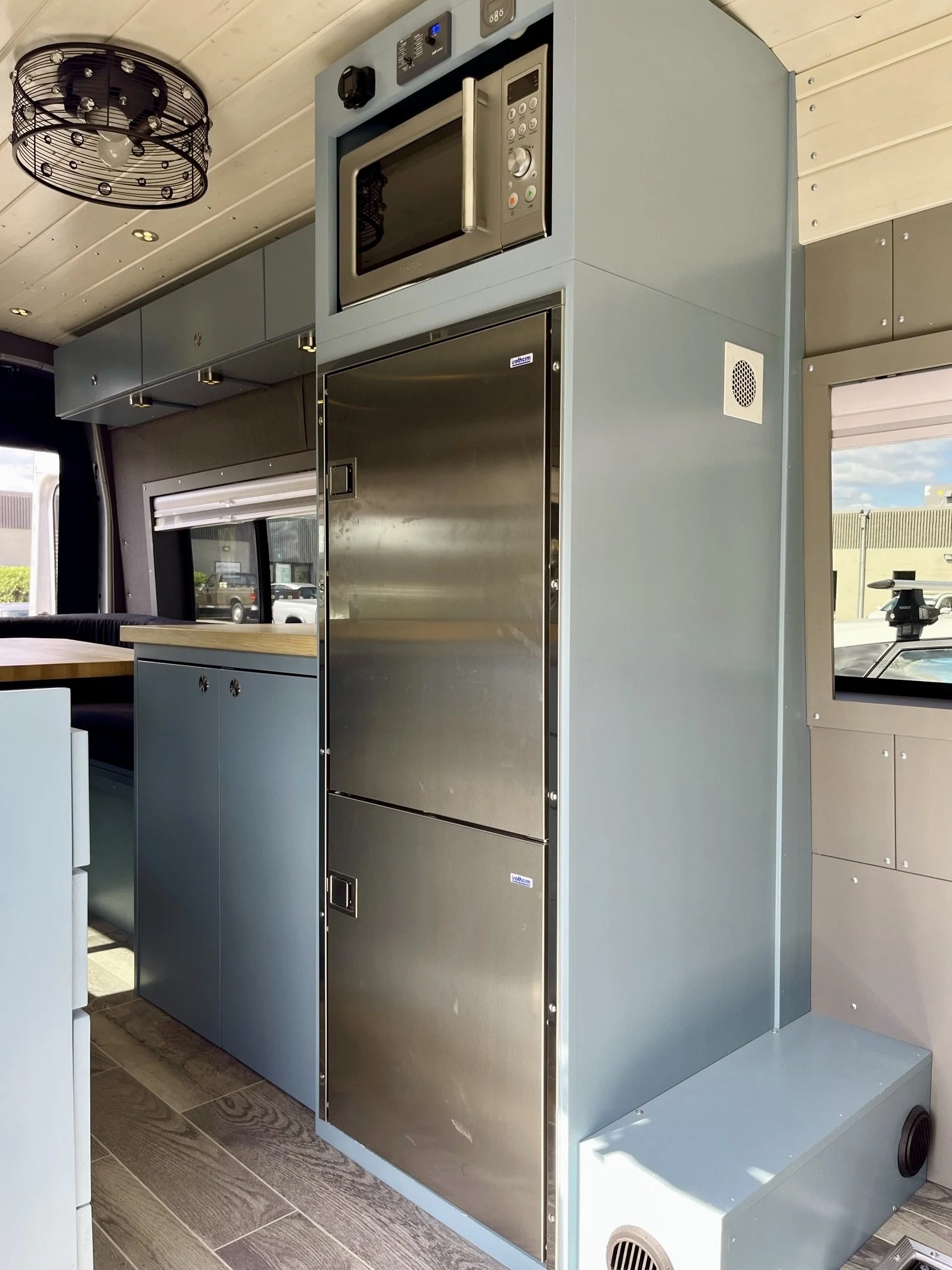 Interior view of a kitchen with a blue cabinet, stainless steel microwave above a stainless steel refrigerator, and a light fixture on a wooden ceiling.