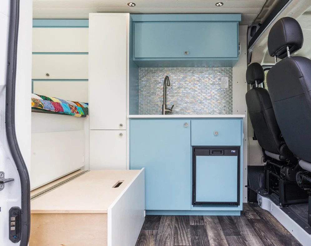 Interior of a van converted into a small living space with a kitchenette, including a sink, fridge, and cabinets, and a sleeping area with a colorful quilt.