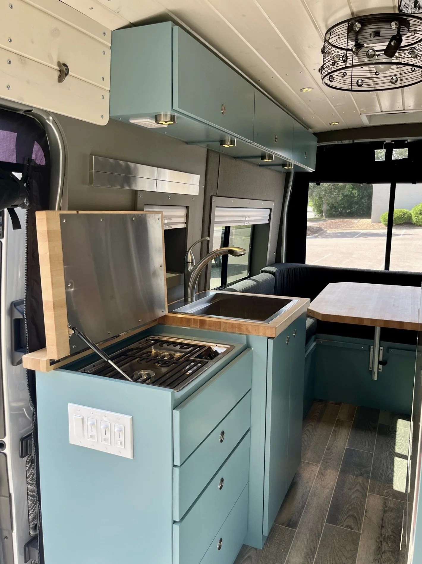 Interior of a compact kitchen area inside a mobile unit or RV, featuring a small cooktop, sink, blue cabinetry, and a window with a view of a parking lot and trees outside.