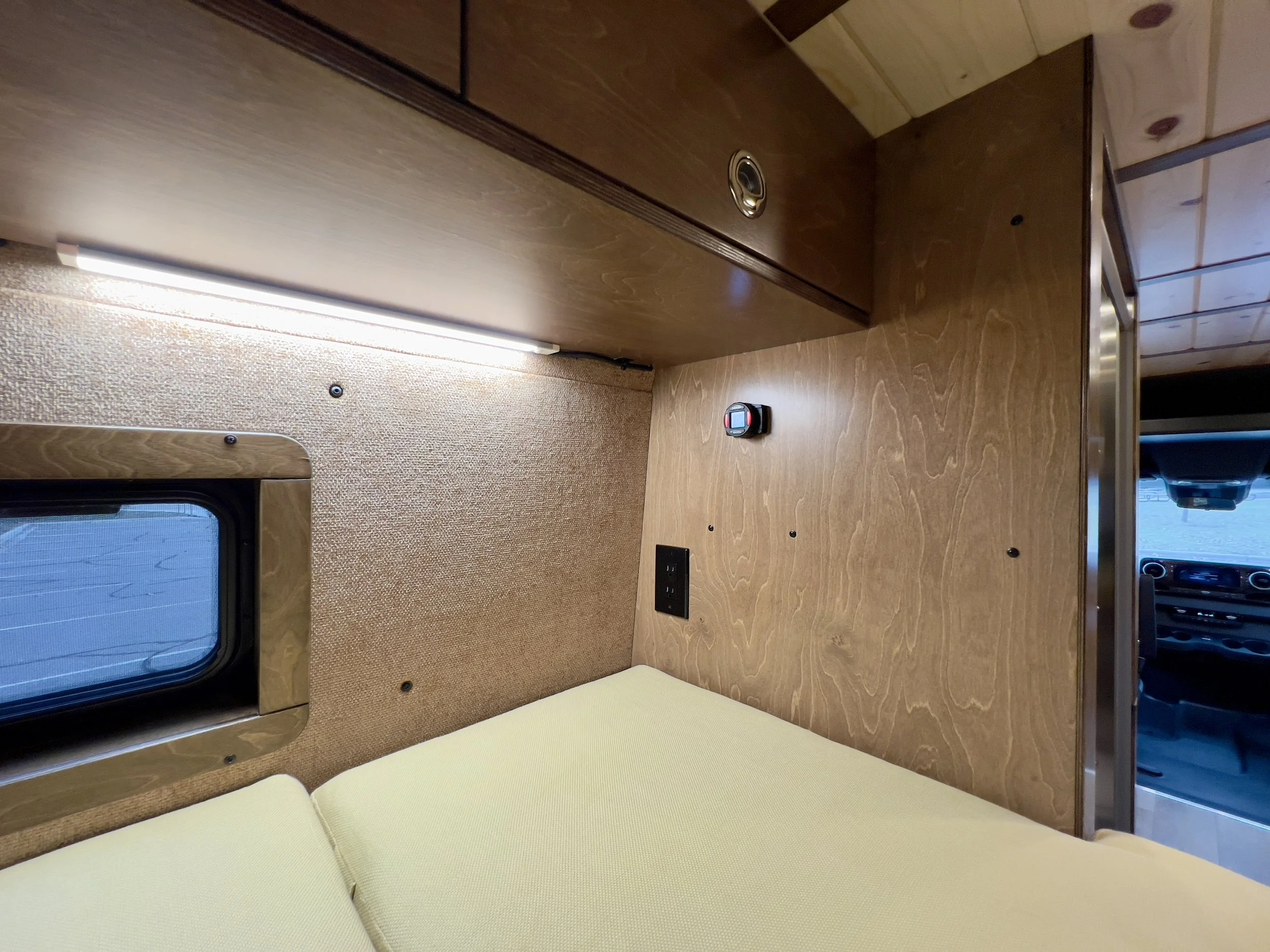 Interior of a small wooden camper with a yellow couch, a window, a power outlet, a small digital clock, and a wood-paneled wall.