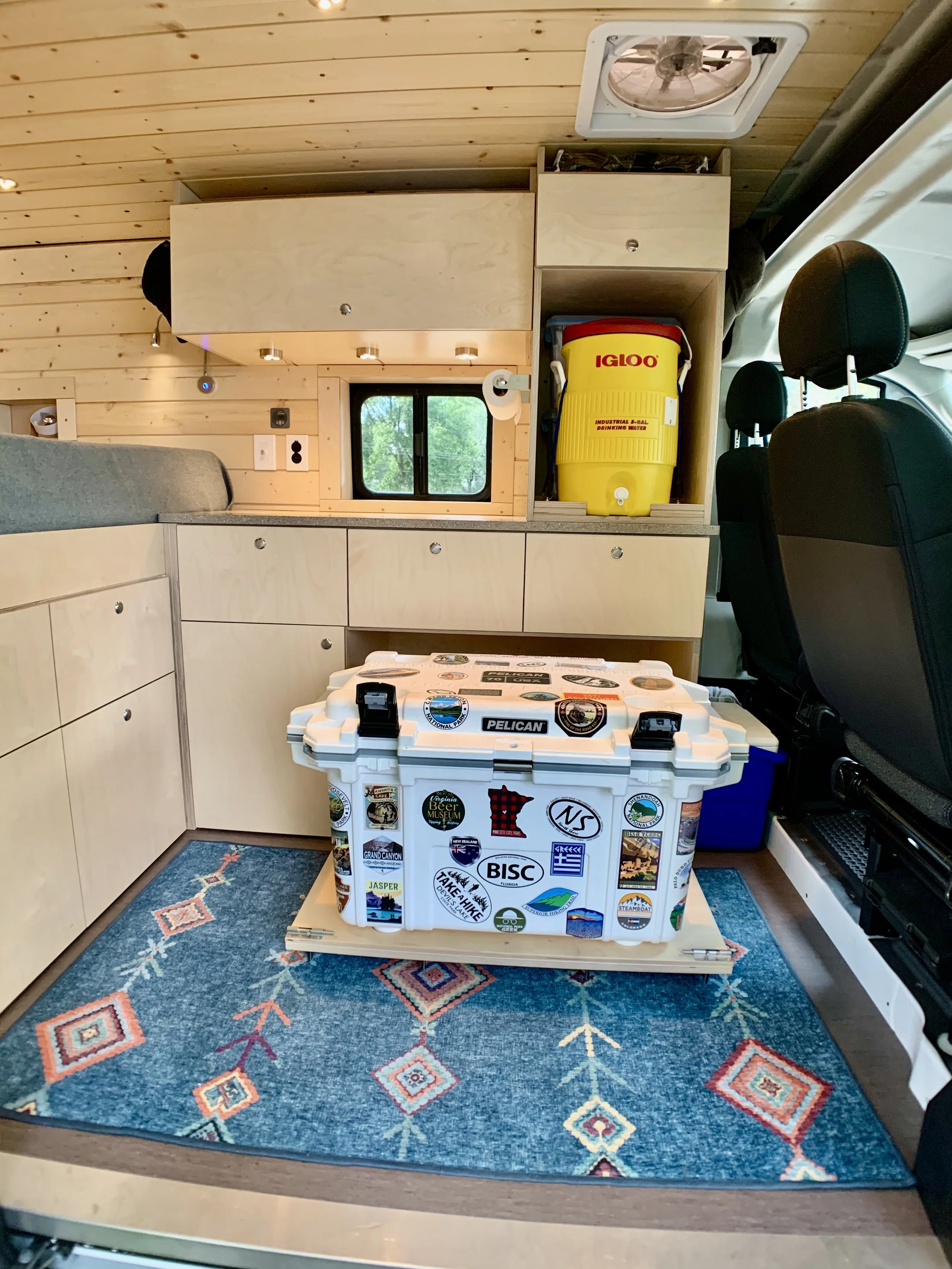 Interior of a van with wooden cabinets, a small window, and a colorful rug on the floor. There is a white cooler decorated with various stickers on the floor, and a yellow Igloo water container on a shelf. The van has a ceiling vent and two front seats.