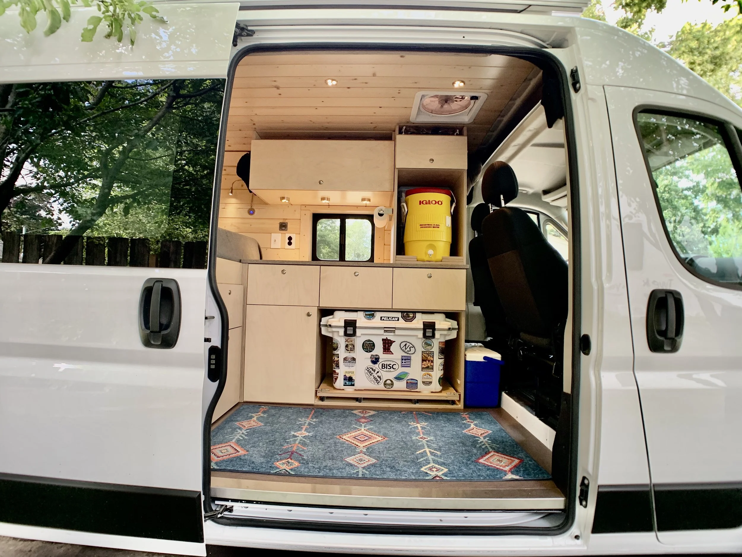 Interior of a camper van converted for sleeping and storage, featuring wooden cabinets, a patterned rug, a cooler, a yellow Igloo cooler, and a window with greenery outside.