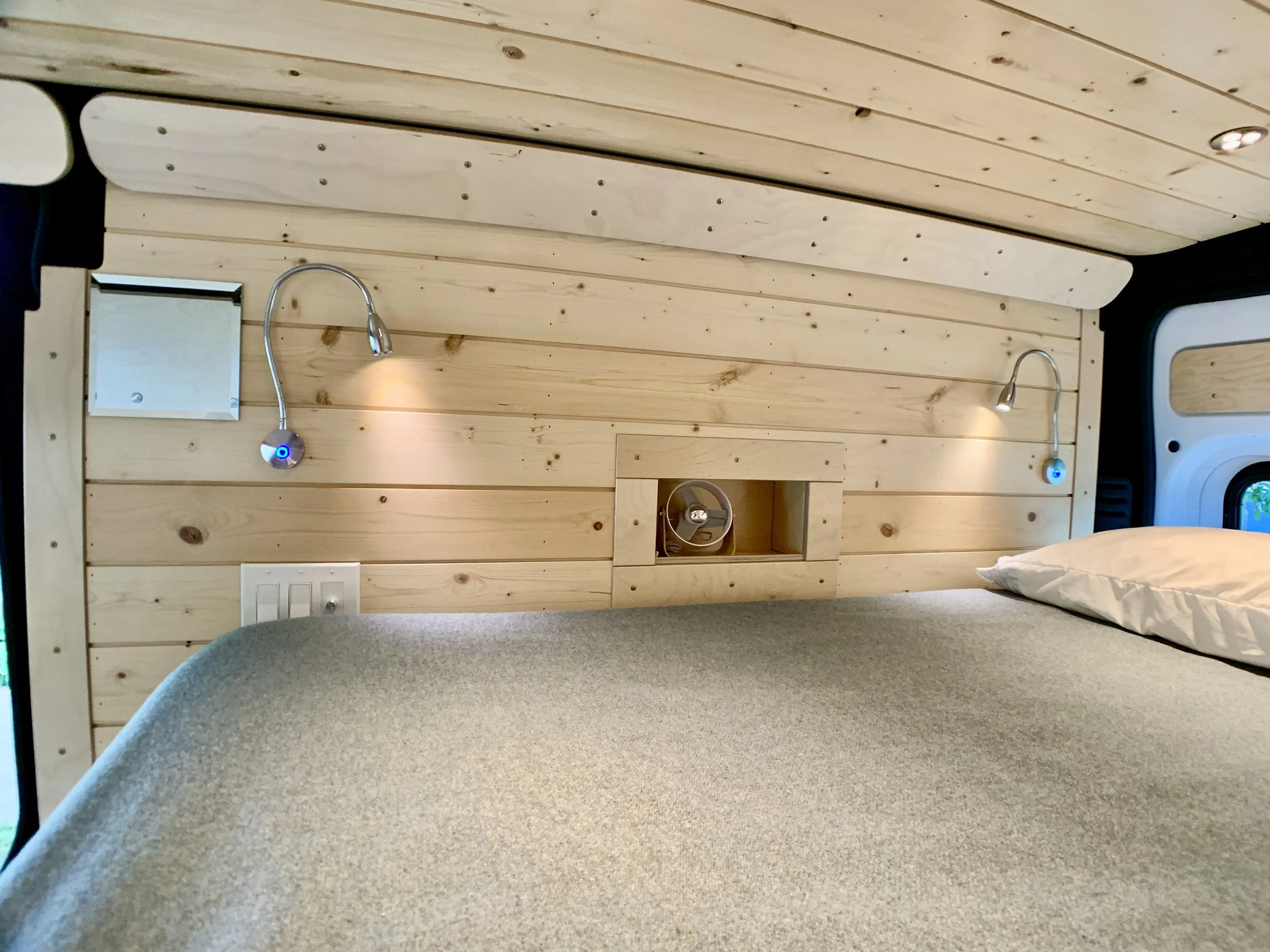 Interior of a wooden camper van with a bed, two reading lights, a small mirror, a window, a fan, and electrical outlets.