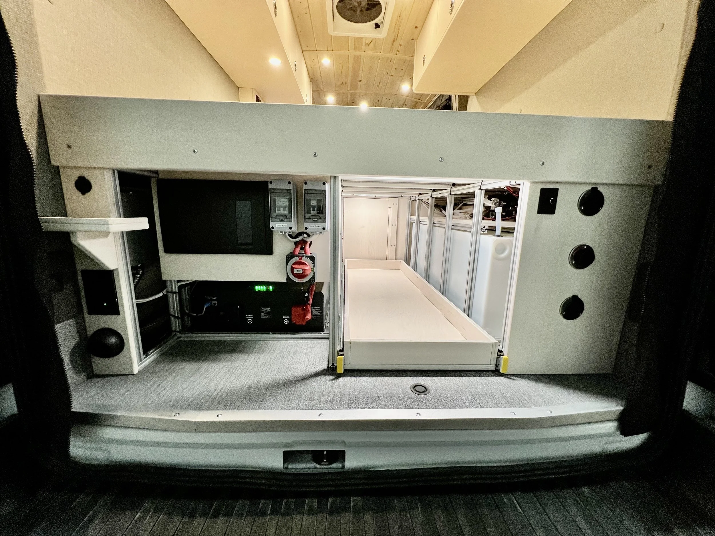 The interior of a van converted into a mobile workspace with electronic equipment, including two monitors, a black power supply or server, and an empty wooden platform or bed area.