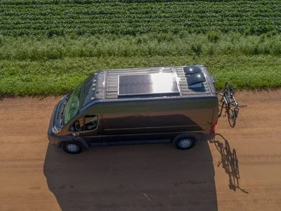 A black van with solar panels on the roof, driving on a dirt road with green fields in the background, and a bicycle attached to the back.