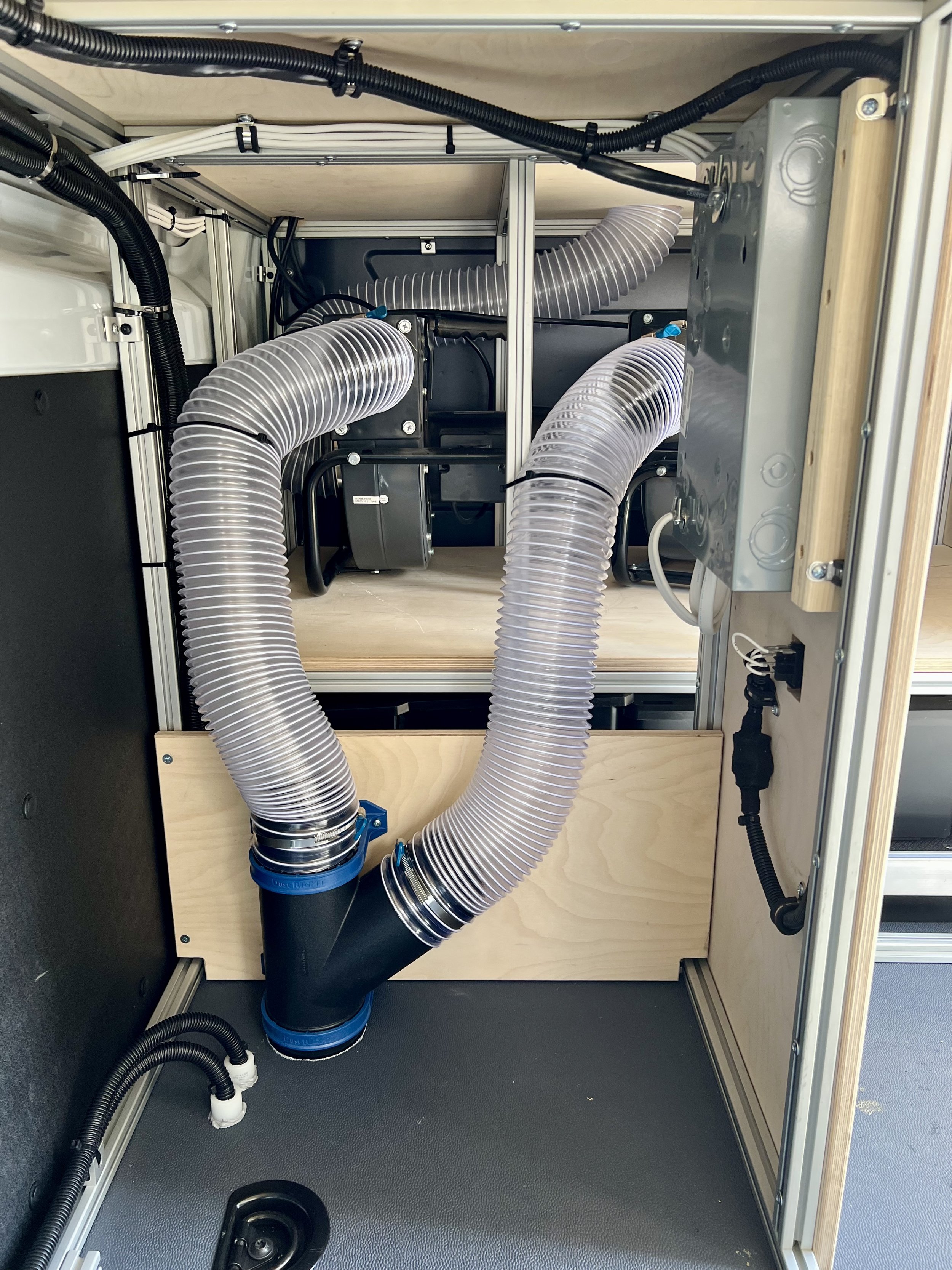 The image shows the interior of a technical equipment or machine enclosure with large silver flexible tubes connected to black fittings. There are also electrical components and wiring attached to the metal frame and wooden panels.