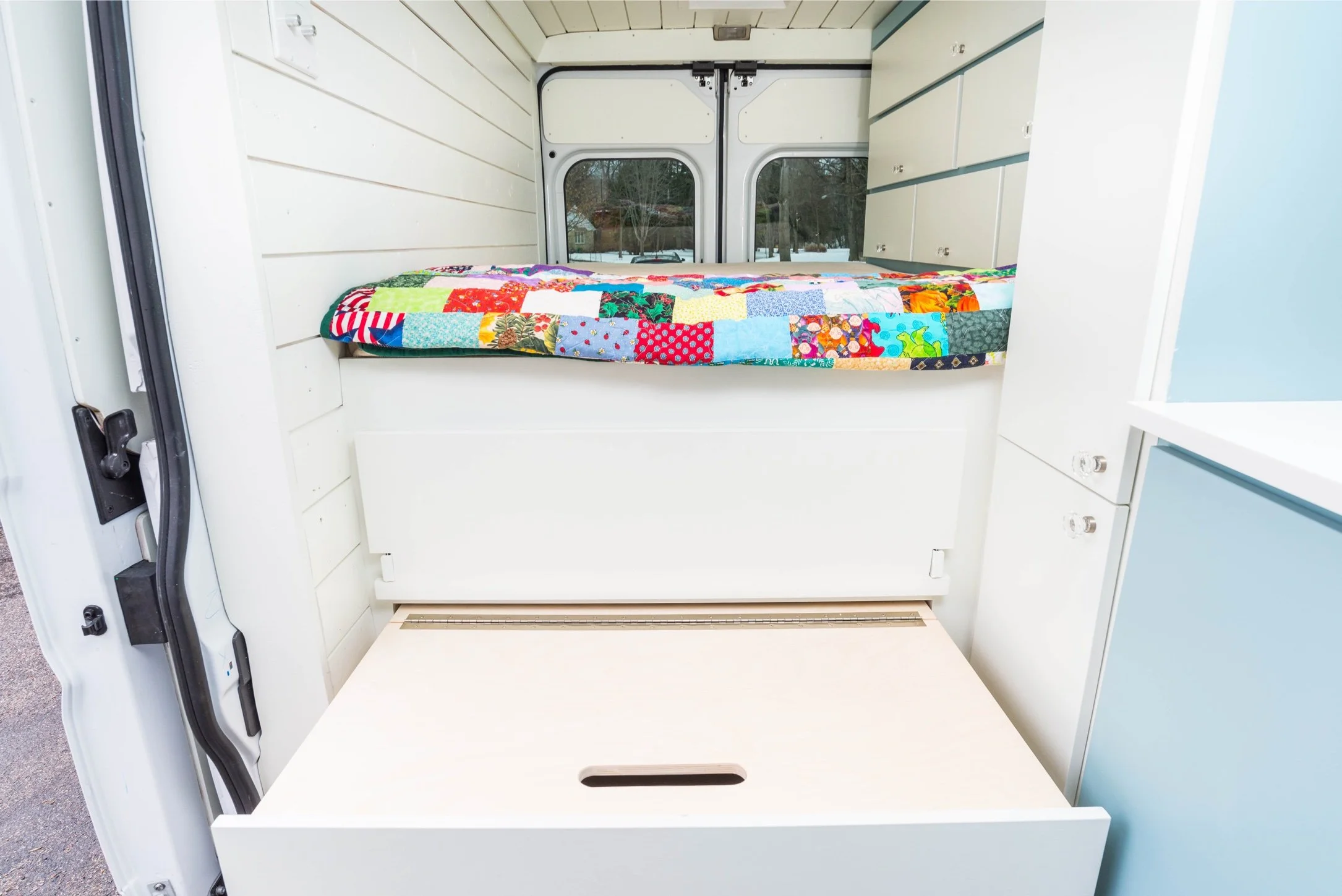 Interior of a camper van with a bed covered by a colorful quilt, and a fold-down table below the bed.