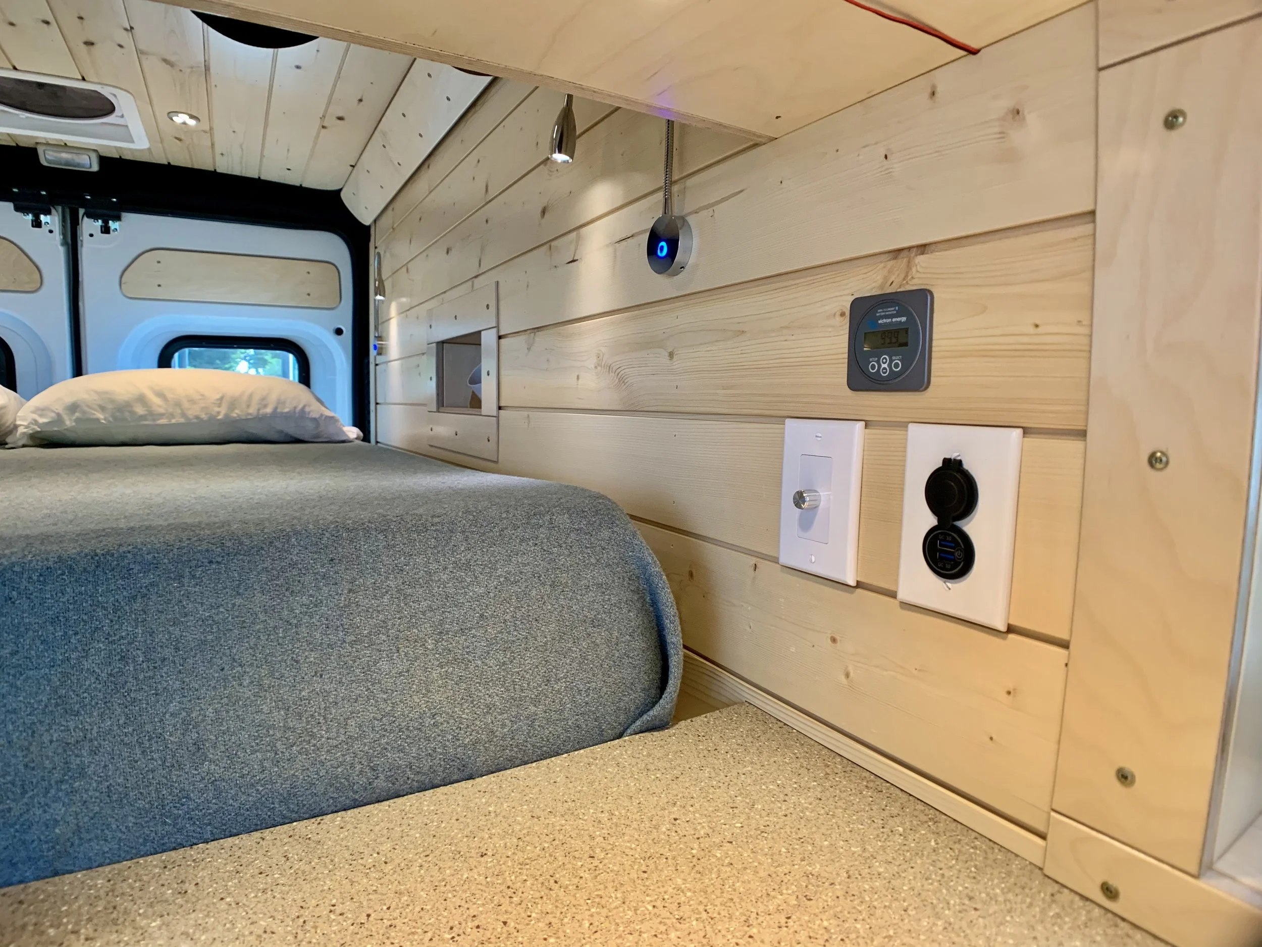Interior of a tiny home or camper with a bed, light wood panel walls, electrical outlets, a control panel, and a small window.