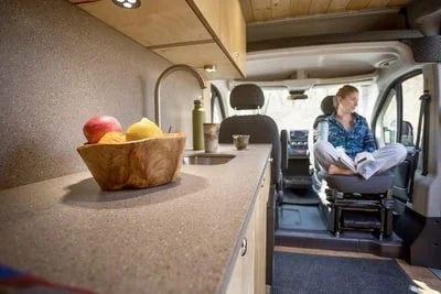 Inside a van with a wood bowl of apples on a kitchen counter, and a person sitting in the driver's seat reading a book.