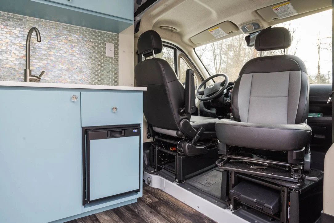 Inside of a converted camper van showing the front seats and a kitchenette with a sink, mini fridge, and blue cabinetry.