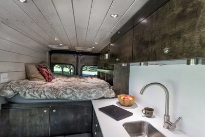 Interior of a camper van with a bed, kitchen counter, sink, and window view.