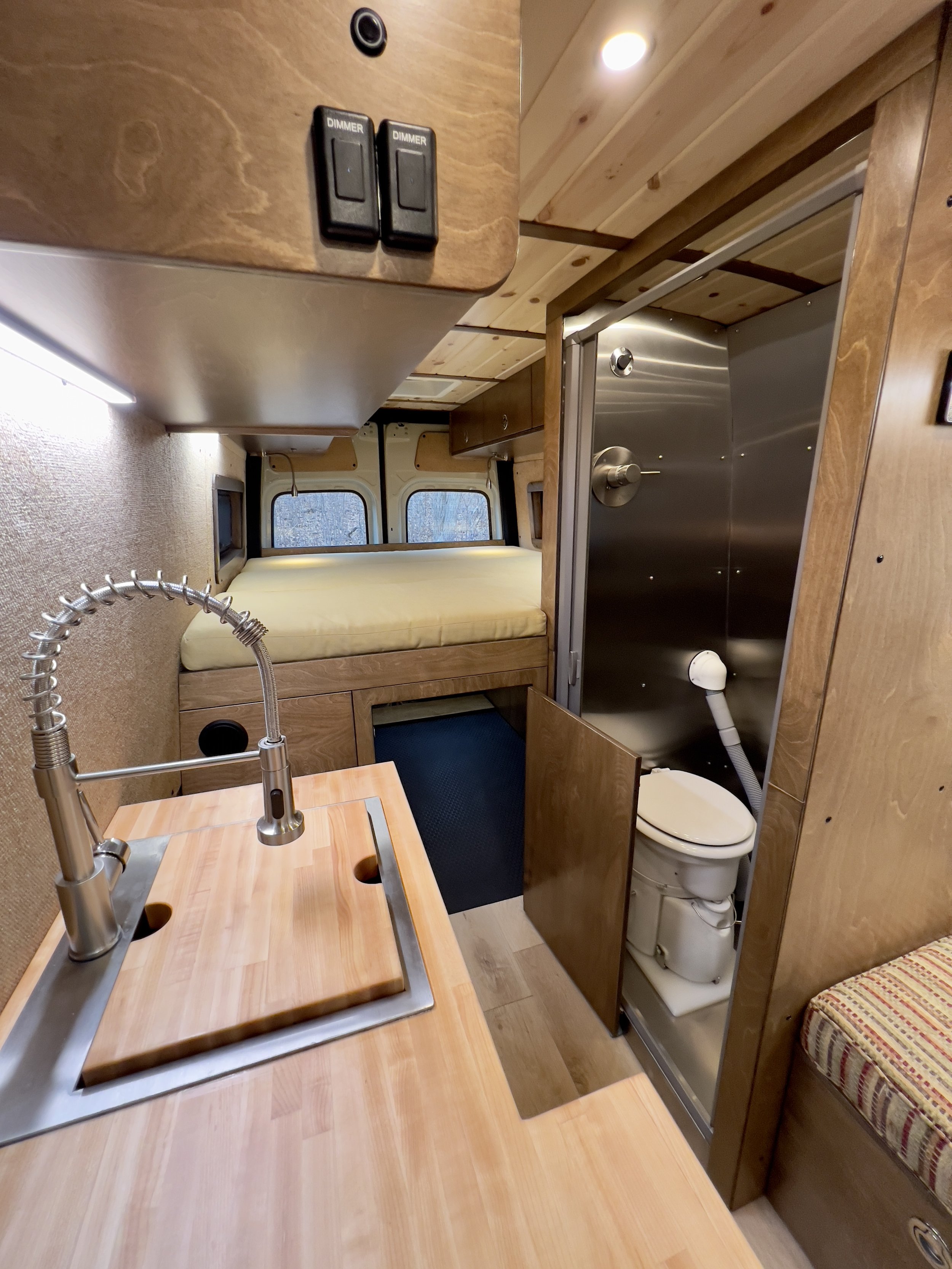 Small RV interior showing a compact kitchen with a wooden countertop and sink on the left, a closed cabinet below, and a small seating area or bed at the back, with windows above. To the right, there's a bathroom with a toilet and metal walls, and on the ceiling, two dimmer switches.