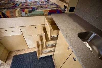 Kitchen with open lower drawers, a sink, and a countertop, with a colorful quilt hanging in the background.