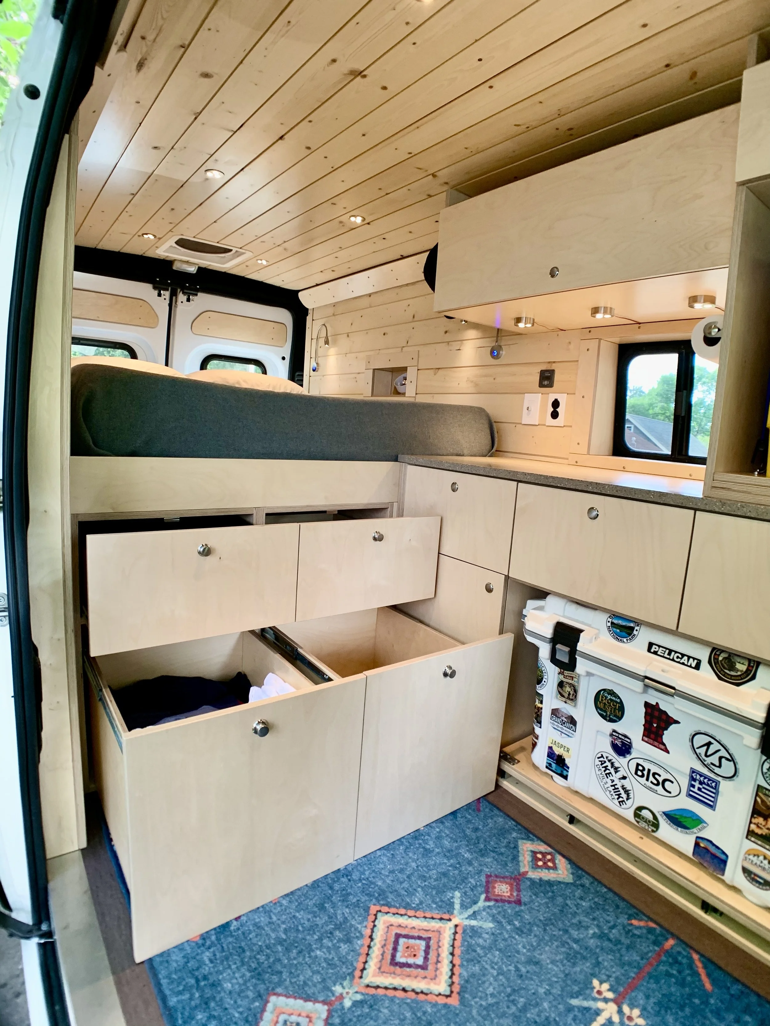 Interior of a converted van with wooden cabinetry, a bed, and a colorful patterned rug on the floor.