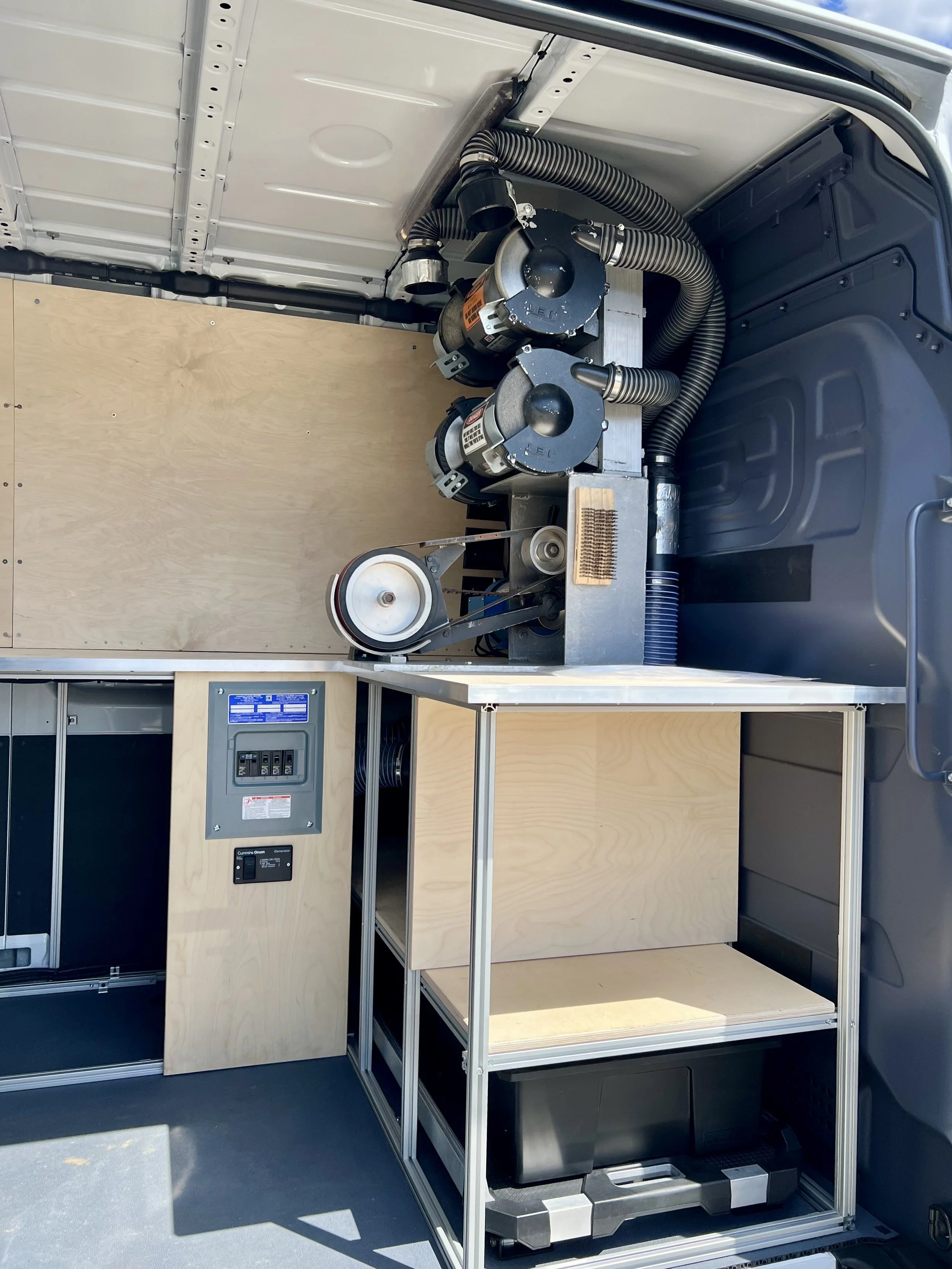 Interior of a vehicle with equipment and wooden shelves, including two black cylindrical motors, flexible hoses, a wheel with a belt, and electrical panels.