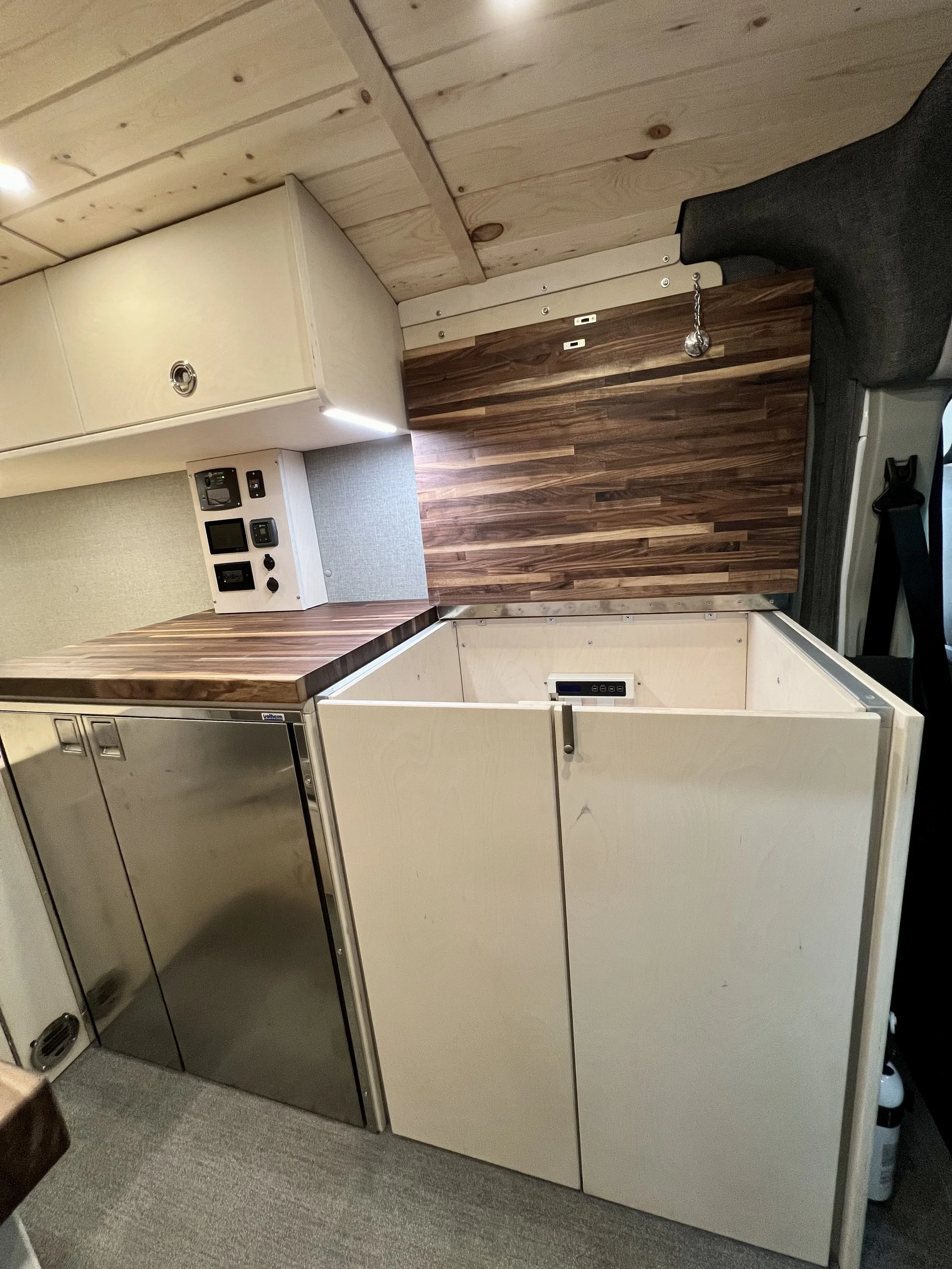 Interior of a compact kitchen with light wood cabinets, a countertop, and a small refrigerator. There are electrical outlets and controls on the wall, and the ceiling is also made of light wood paneling.