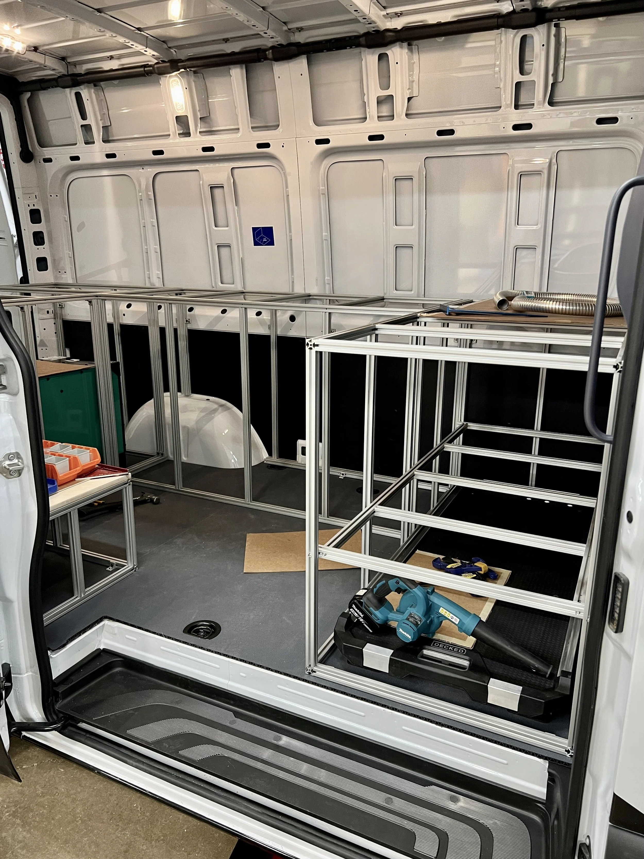 The interior of a work van in the process of being set up with a metal framework for storage or equipment, with a power tool on the floor, some pipes, and small storage containers.