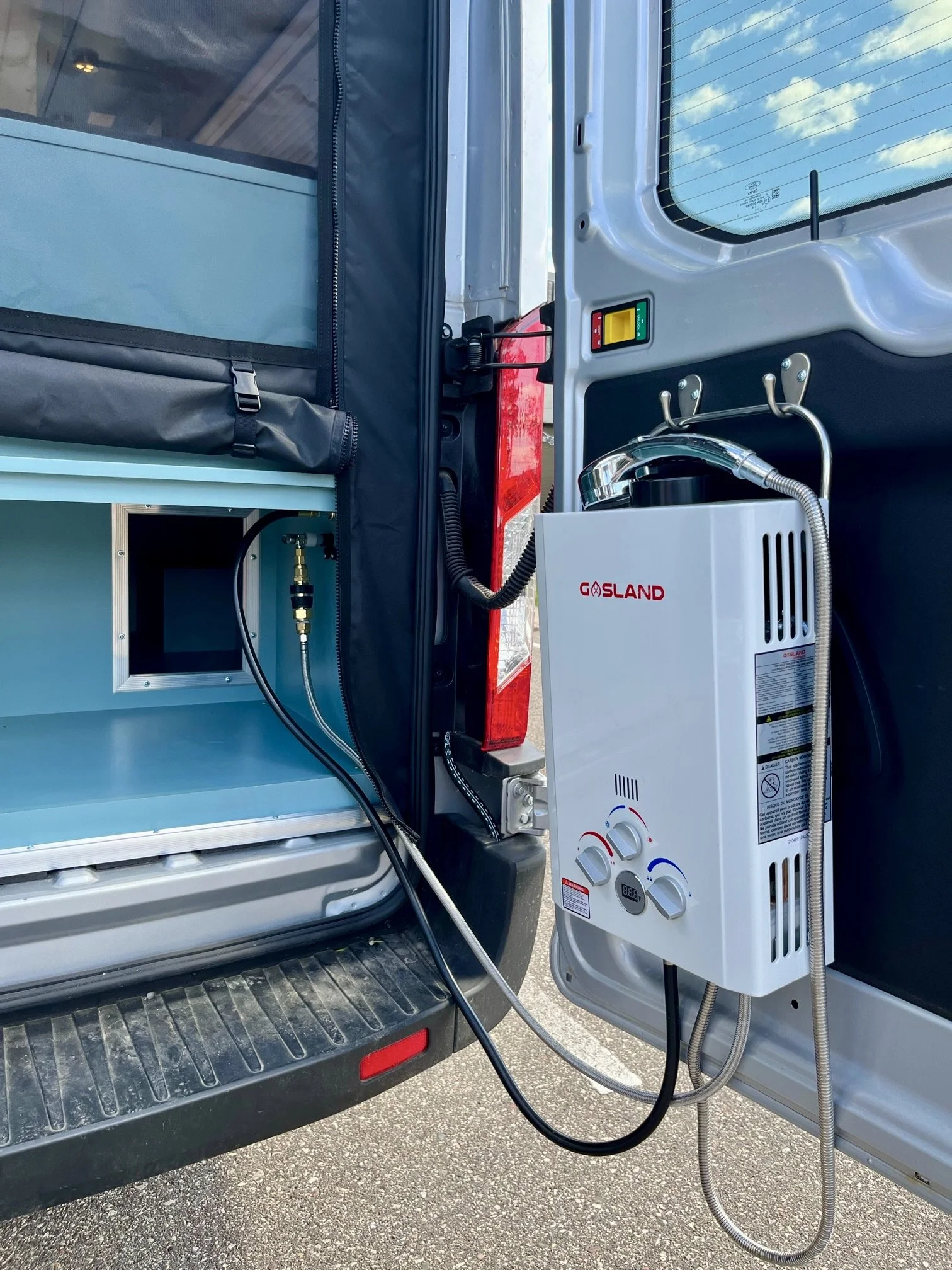 A portable white water heater labeled GØSLAND is mounted inside the rear of a vehicle, with hoses connected for supplying hot water.