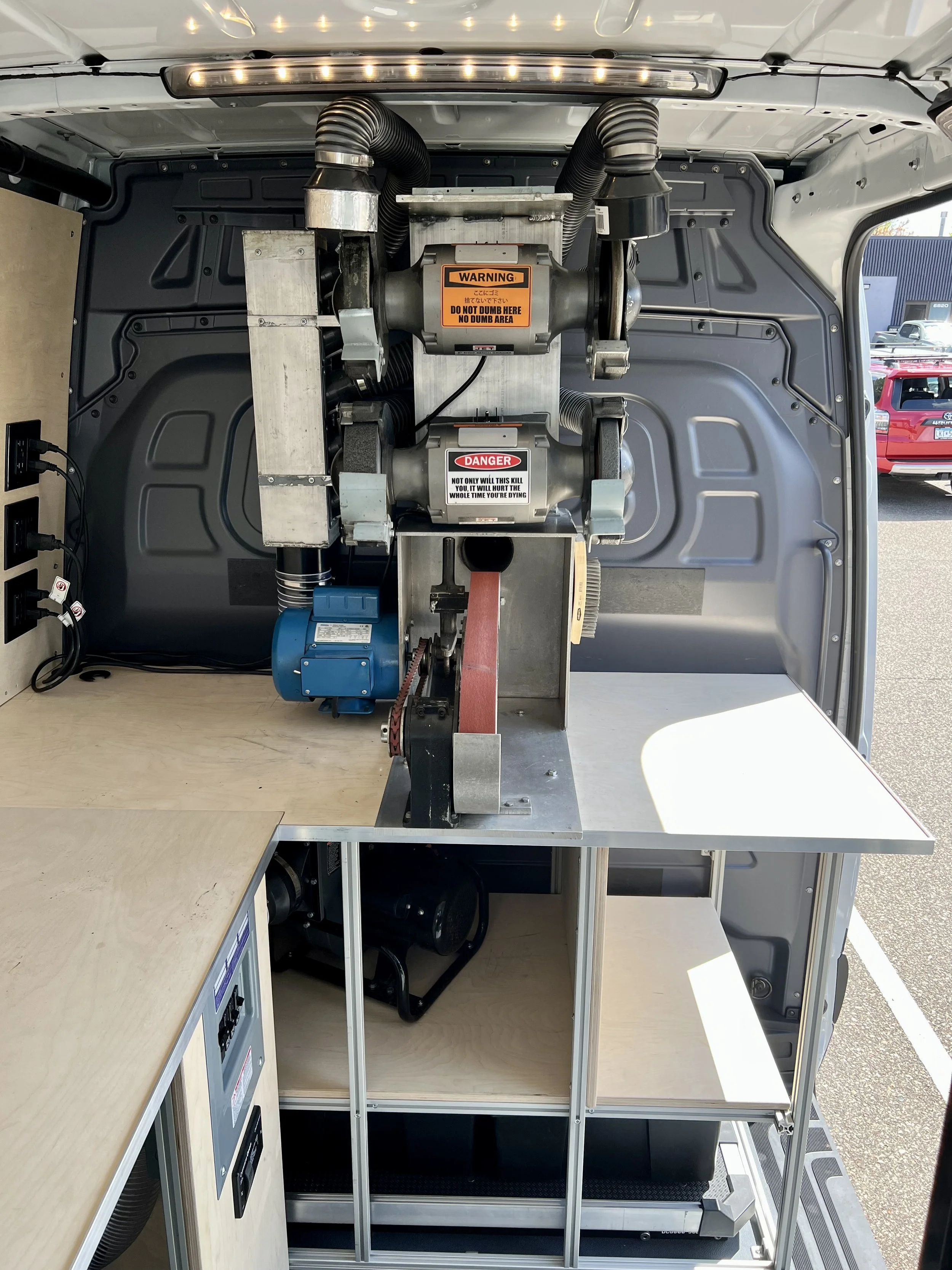 The interior of a mobile workshop van with a large industrial machine, featuring warning and danger labels, mounted on a metal stand, with wooden work surfaces and electrical outlets, set up for technical work or manufacturing activities.