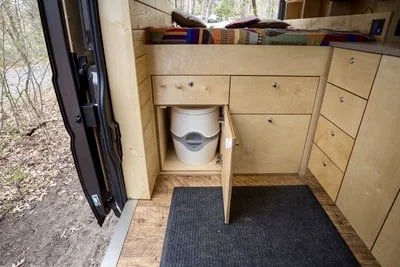Interior of a tiny house or camper showing a toilet inside a wooden cabinet with open door, next to a door leading outside, with a bed and drawers nearby.