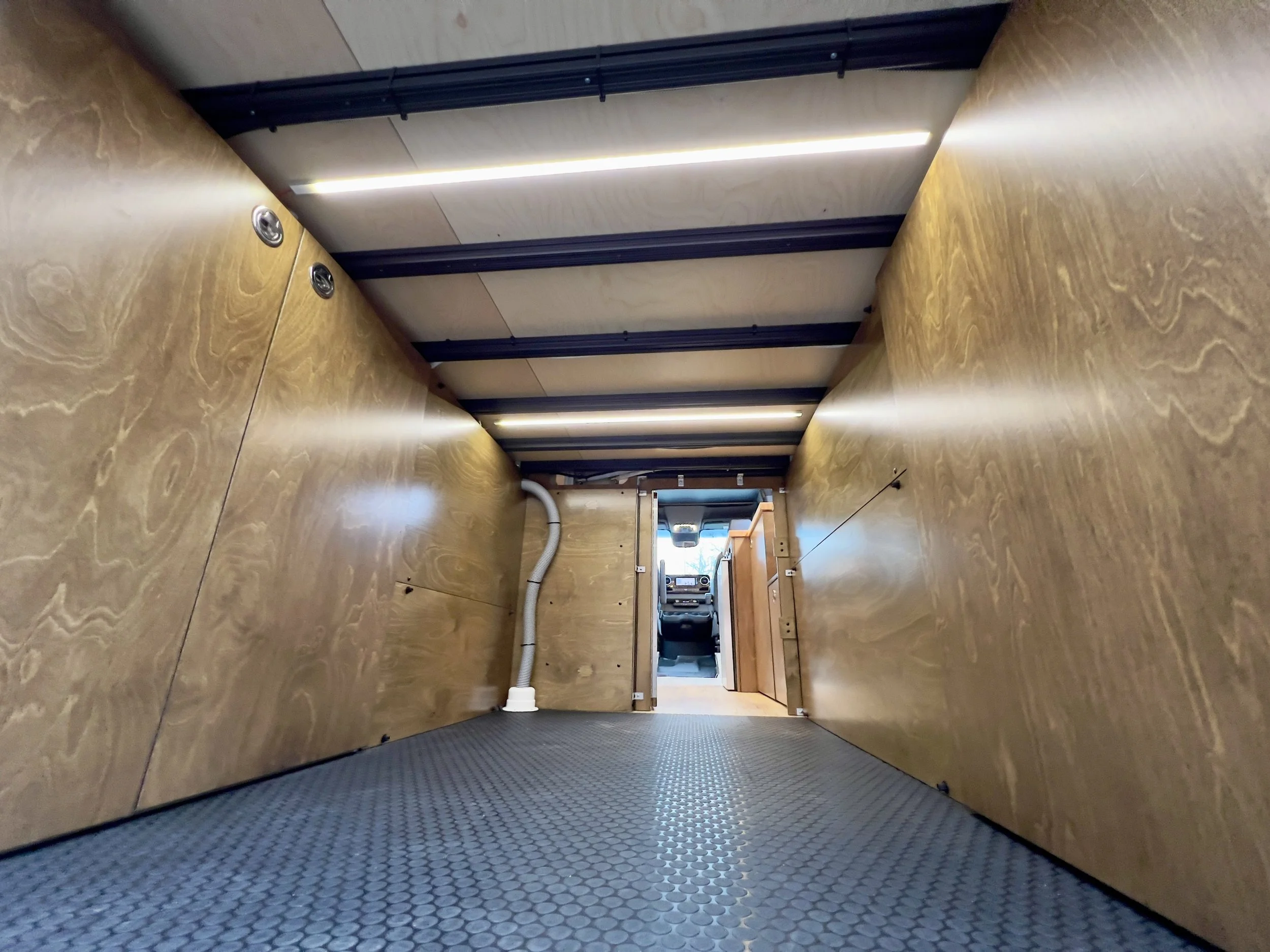 View of an empty cargo area with wooden walls, a metal floor, and a partially visible dashboard at the front, likely taken from inside a delivery van or truck.