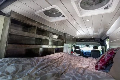 Interior of a camper van with a bed on the left side, stored shelving, a kitchen sink, and a view of the driving area in the background.