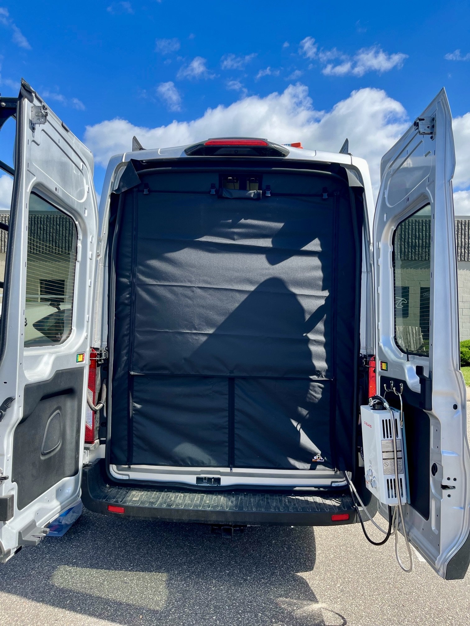 The back of a white service van with doors open, showing a large black insulated storage unit with a machine attached on the right side, under a blue sky with clouds.
