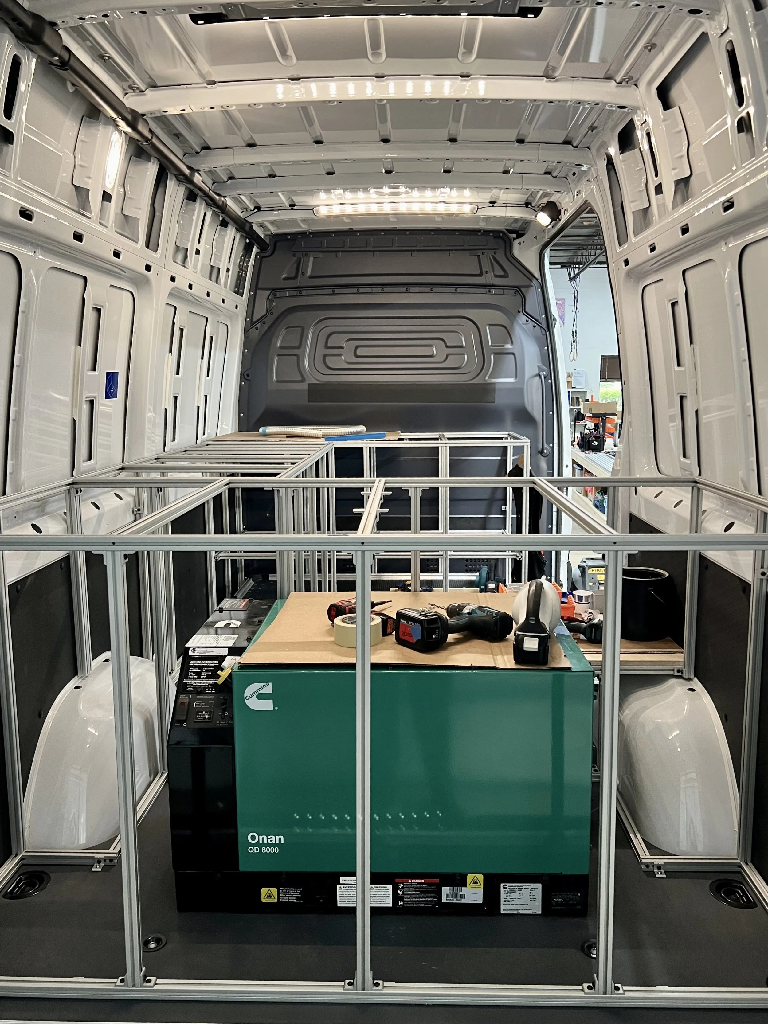 Interior of a work van with unfinished metal framing, tools, and a green Onan QD 8000 generator on a workbench.