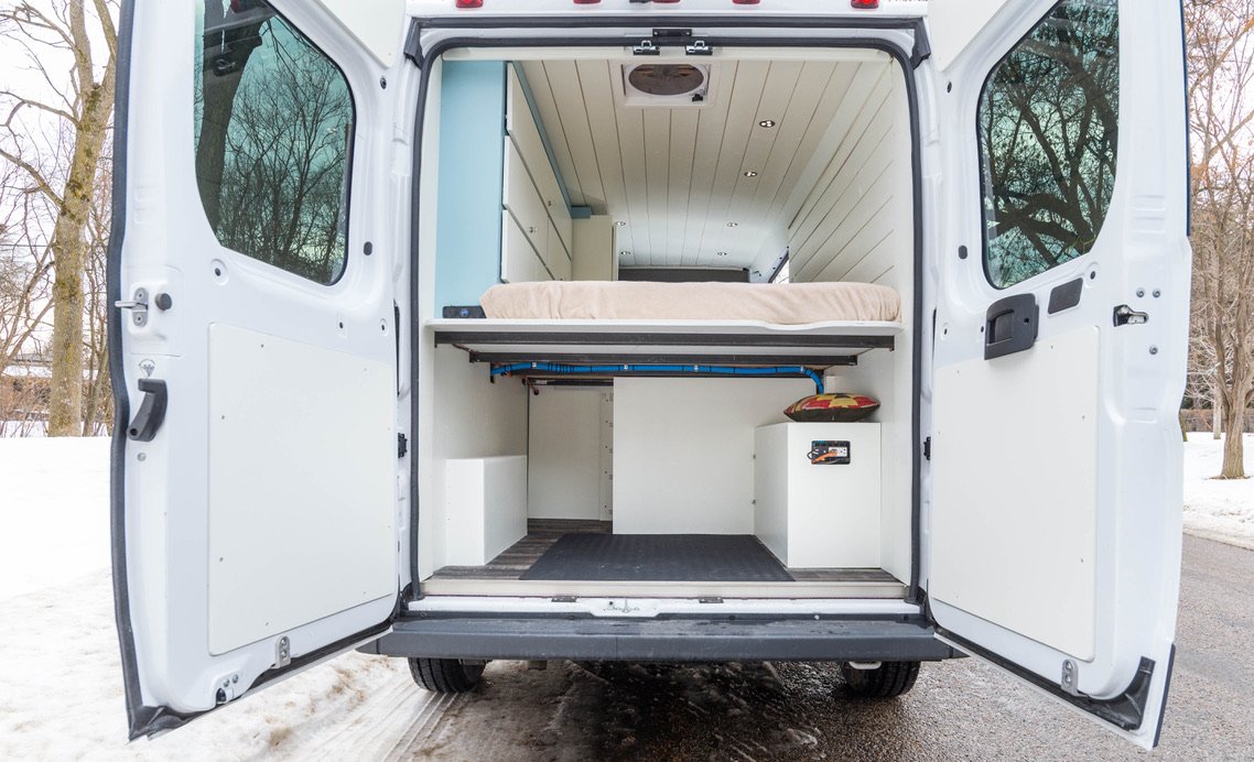 Interior of a converted camper van with a bed and storage cabinets, parked outdoors on a snowy day.