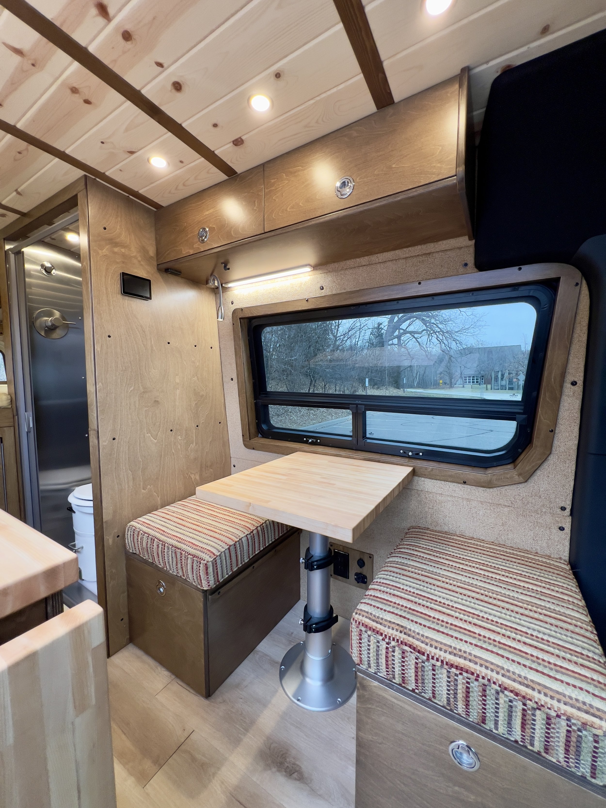 Interior of a small wooden camper or van with a dinette table flanked by two cushioned benches with striped fabric, a window showing trees outside, overhead storage cabinets, and wood-paneled walls and ceiling.