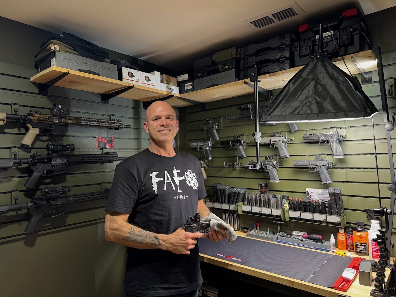 A man smiling in a gun workshop, holding a handgun, surrounded by firearms, gun accessories, and tools on workbench and shelves.