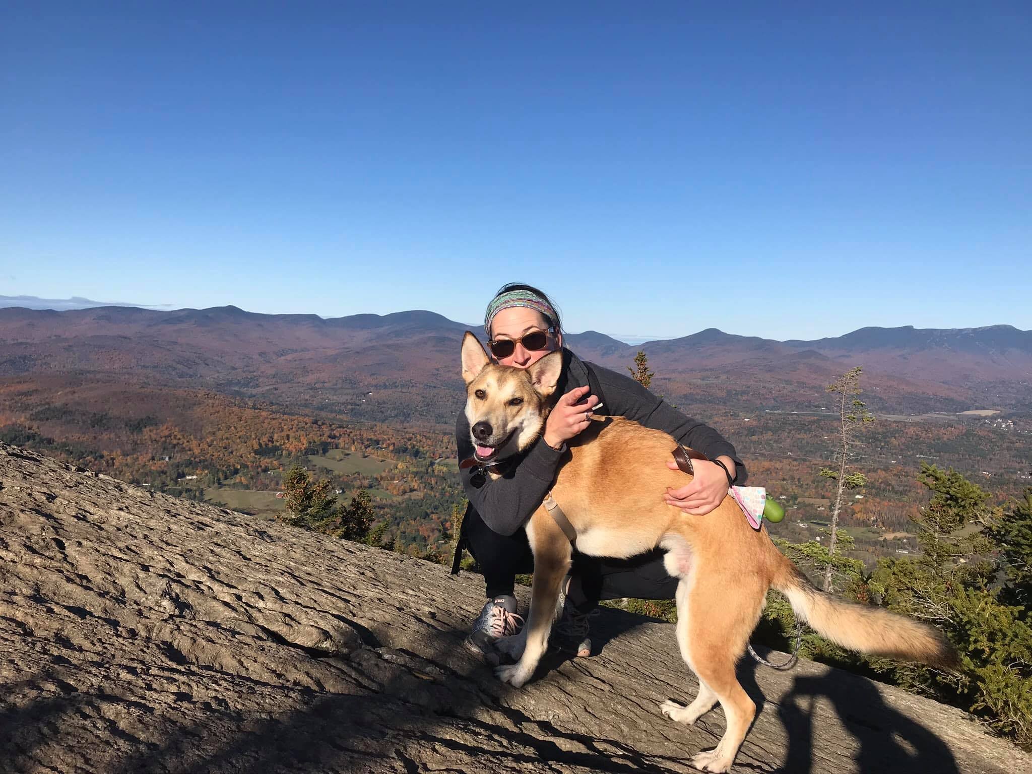 A woman hugging a tan and white dog on a mountain with a scenic view of mountains and trees in the background.