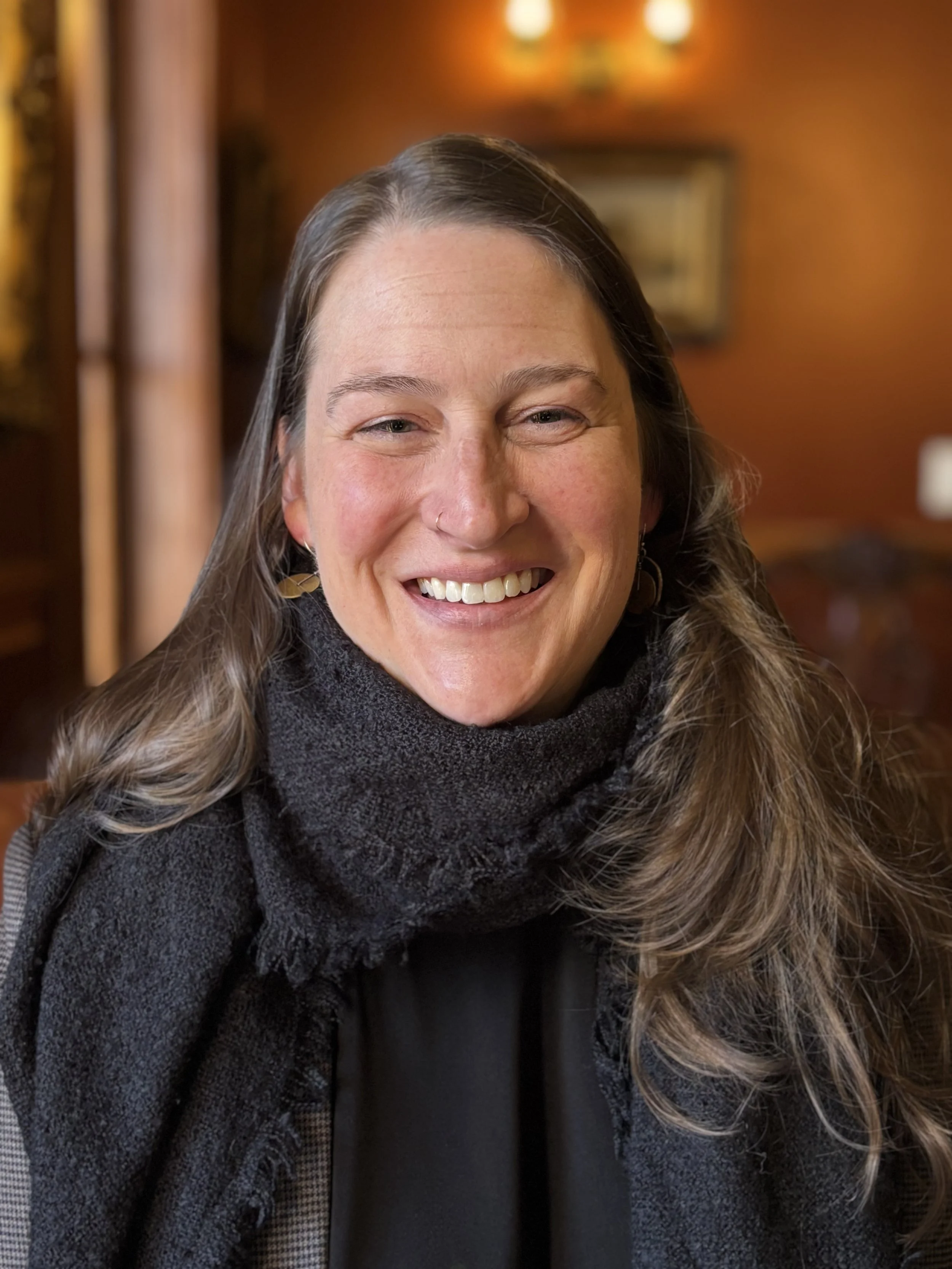 Smiling woman with long brown hair and earrings, wearing a black scarf and coat, indoors with warm lighting and blurred background.