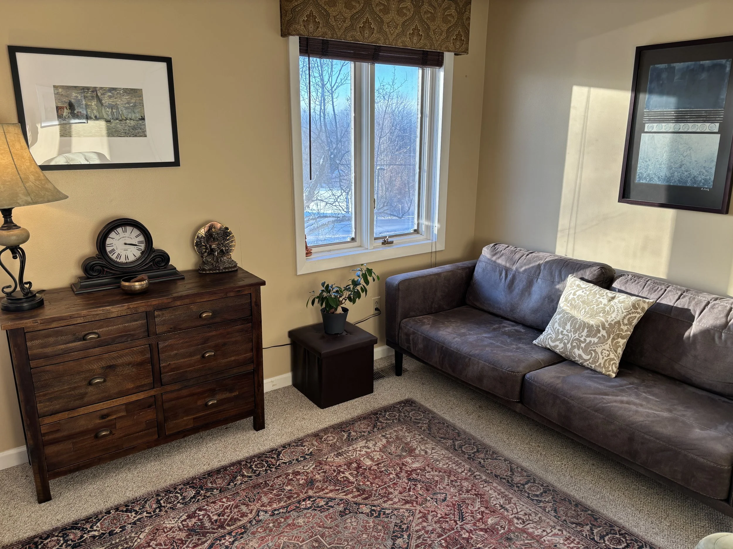Office with beige walls, a window showing snowy outdoors, a dark brown sofa with a decorative pillow, a wooden dresser with a clock, lamp, and sculpture, a potted plant, framed artwork, and a patterned area rug.