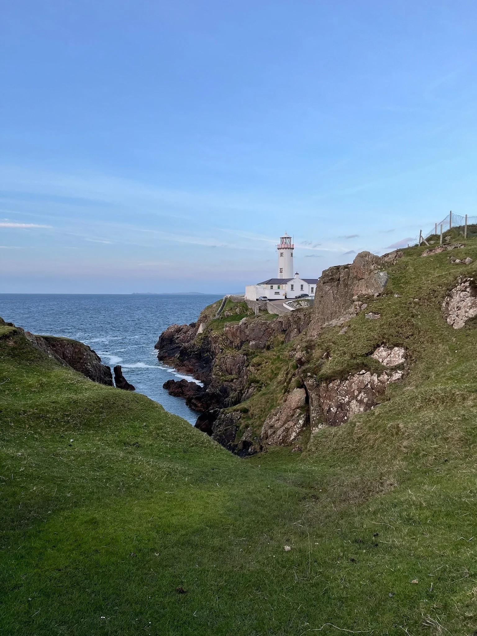 Scenic view of a lighthouse on a rocky coastline with green grass and a calm ocean in the background under a clear sky.