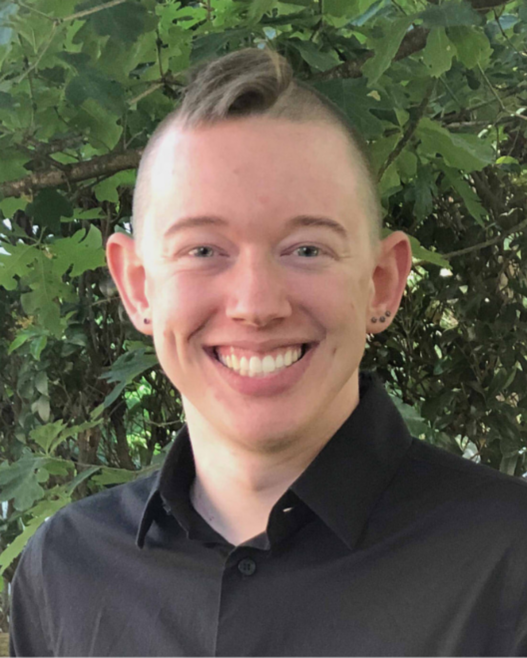 A young man with short, styled hair and earrings, smiling in front of green leafy bushes, wearing a black shirt.